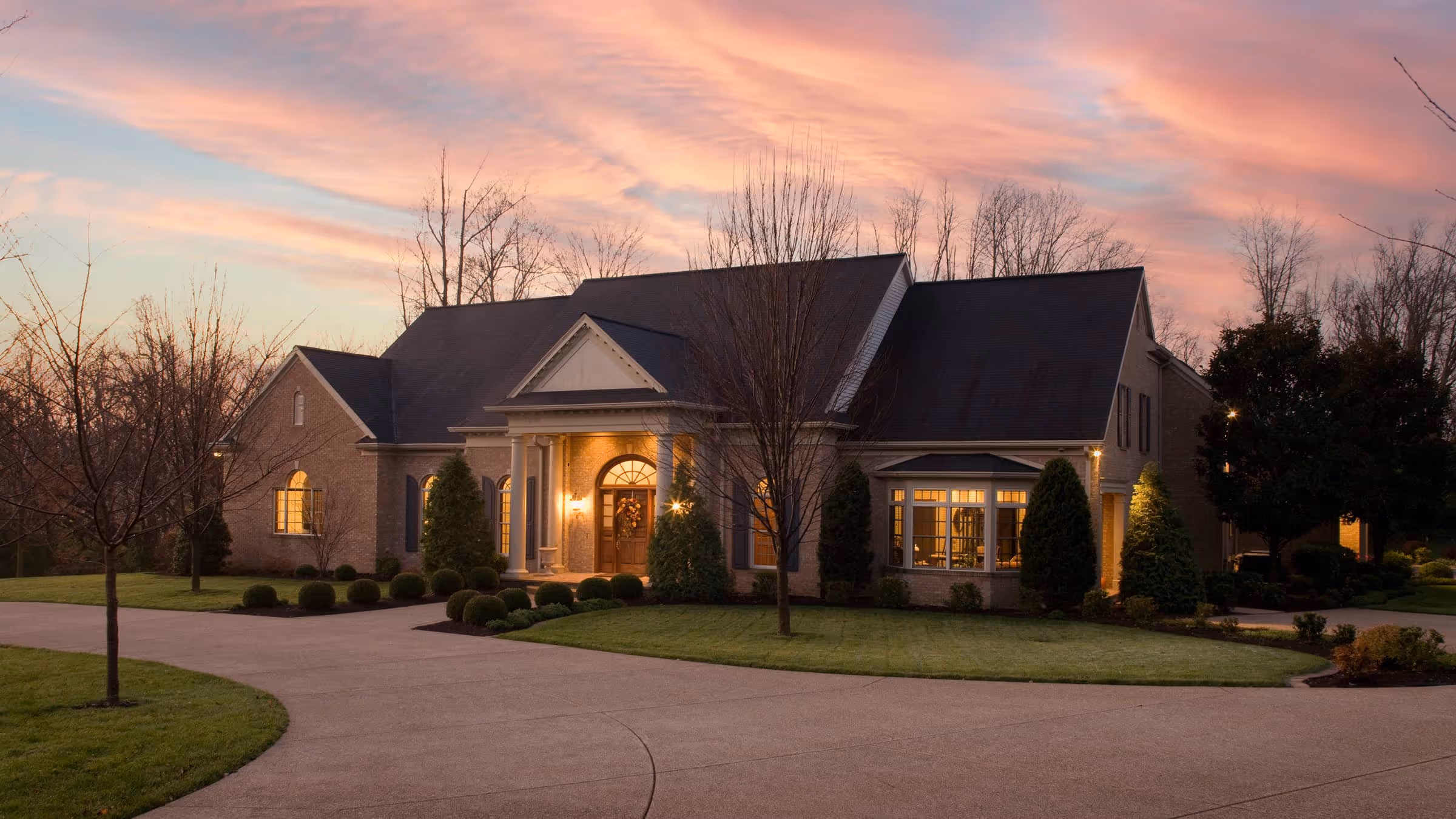 Large brick Federal style house with lit windows and columns at the entrance during sunset with a pink sky.
