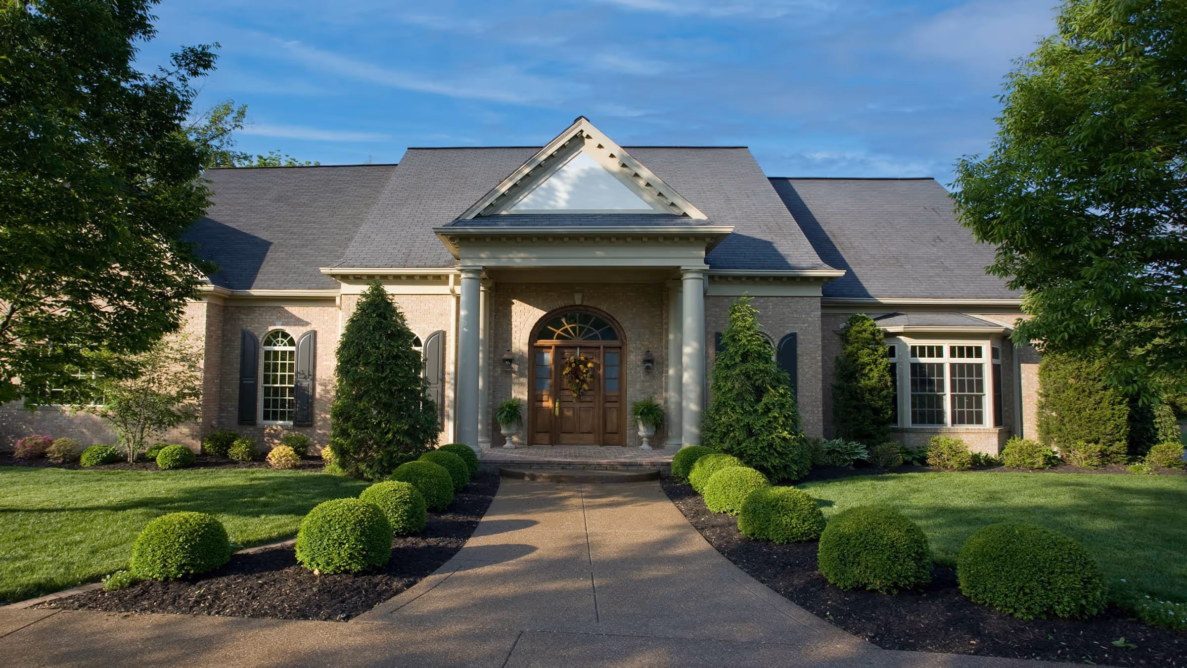 Federal style house with a gray roof, wooden double front doors, and a pathway lined with trimmed green bushes and trees.
