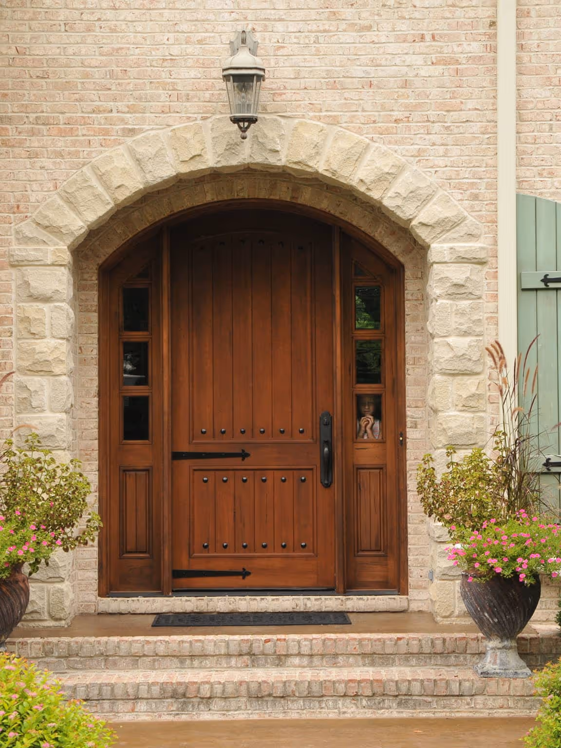 Arched wooden front door set in a stone brick wall with two side windows, stone steps leading up, and large planters with pink flowers on either side.
