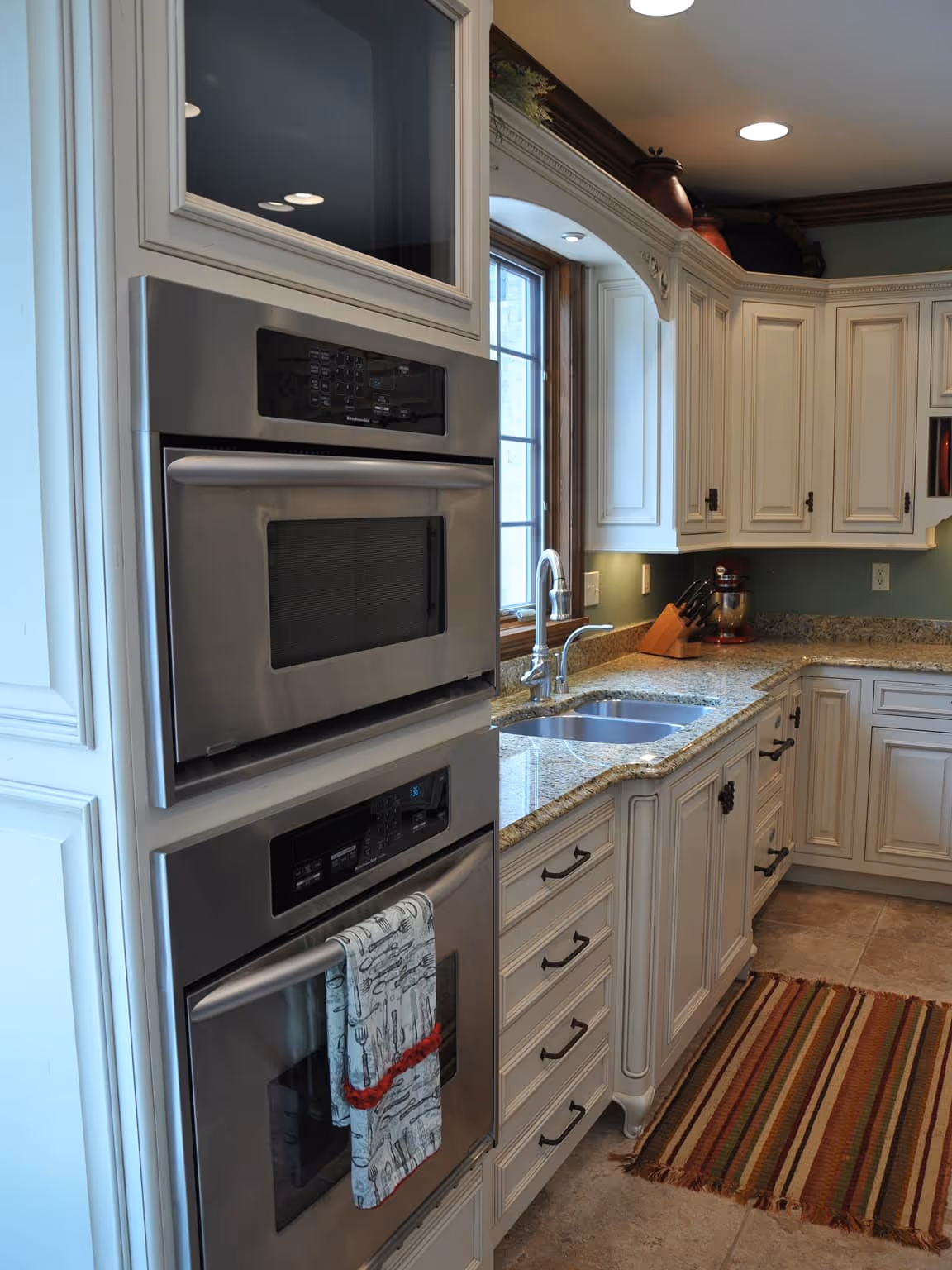 Modern kitchen with double built-in stainless steel ovens, white cabinets, granite countertops, a window above a double sink, and a striped rug on tiled floor.