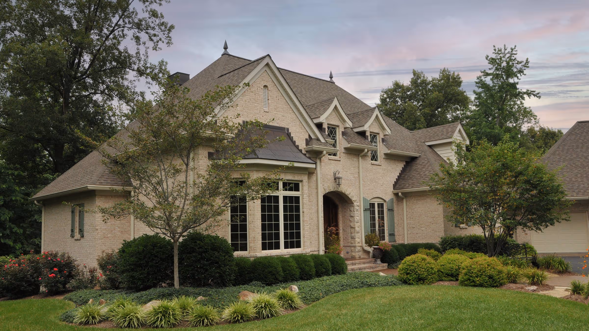 Brick French Eclectic style house with steep gray roof, large windows, and manicured front yard with bushes and trees.