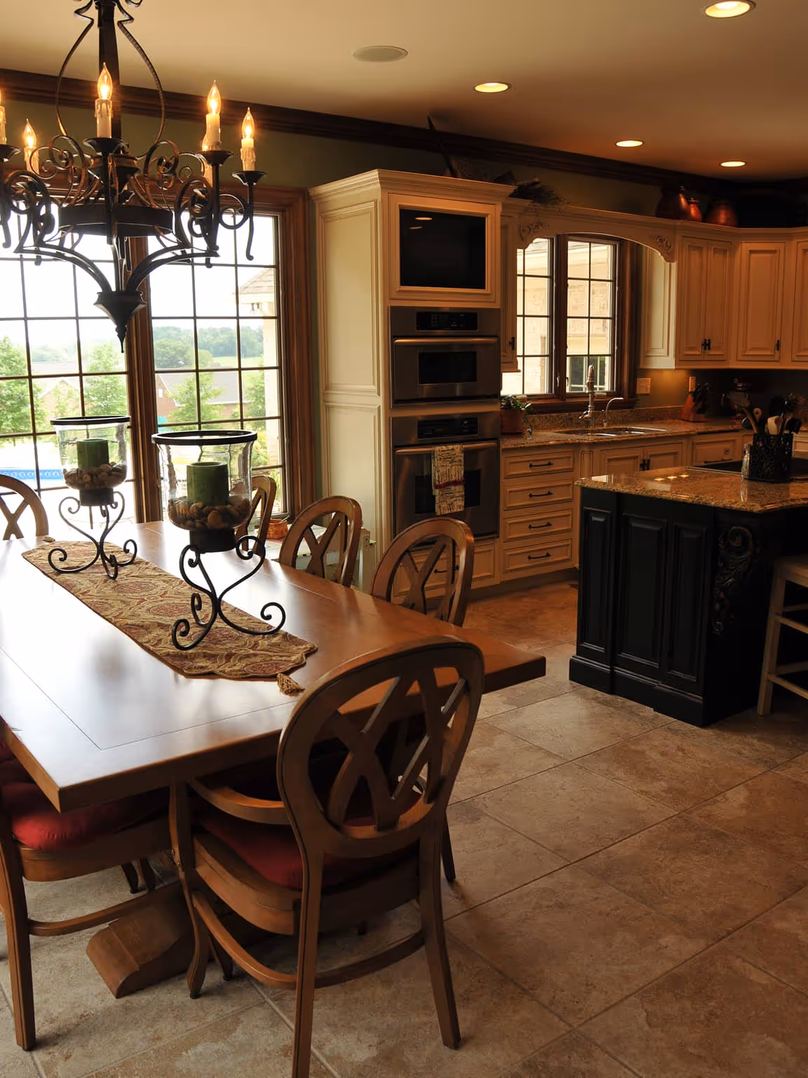 Warm kitchen and dining area with wooden table, decorative candles, chairs with red cushions, granite countertops, and large windows.