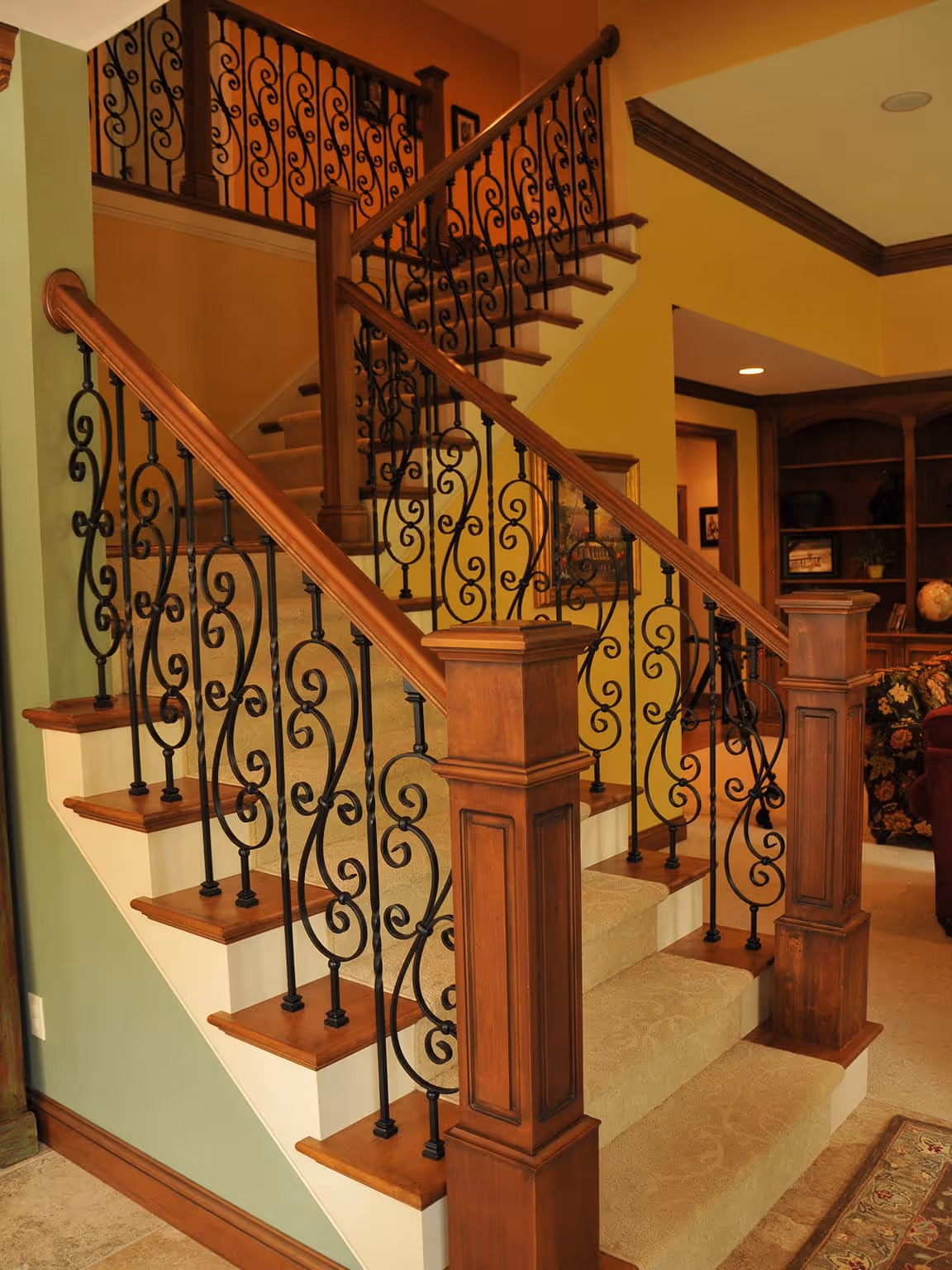 Indoor wooden staircase with beige carpet and decorative black wrought iron railings in a warmly lit living room.