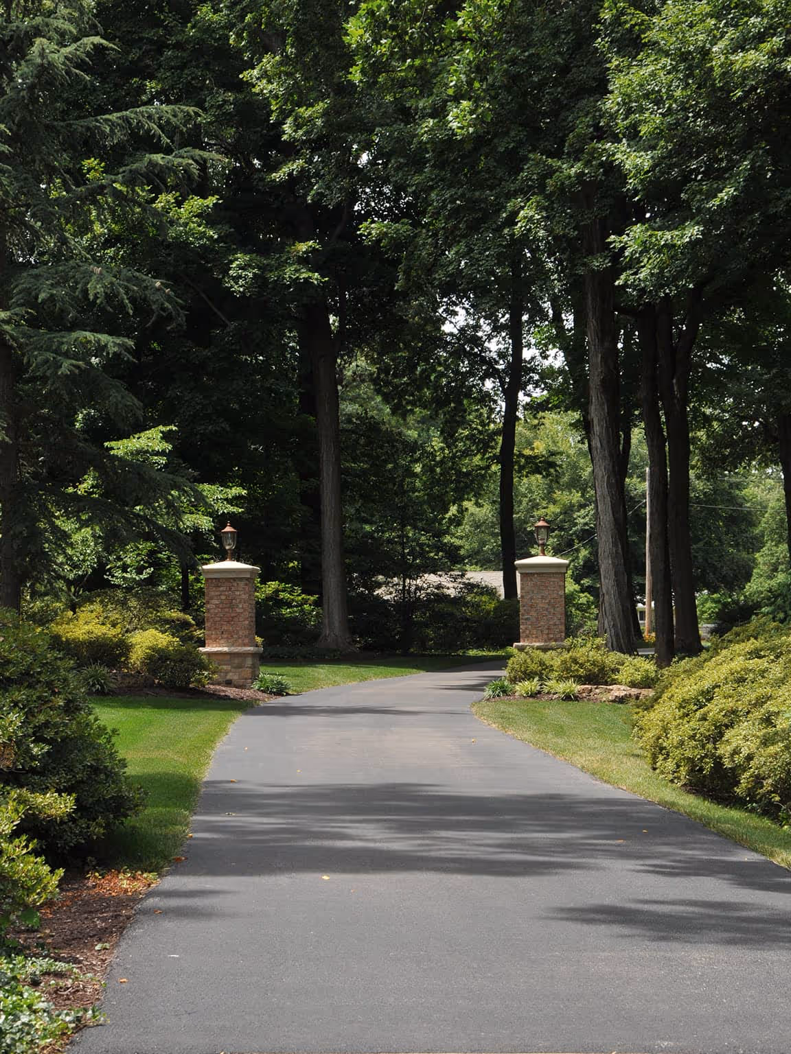 Curved driveway flanked by brick pillars with lanterns, surrounded by green bushes and tall trees.