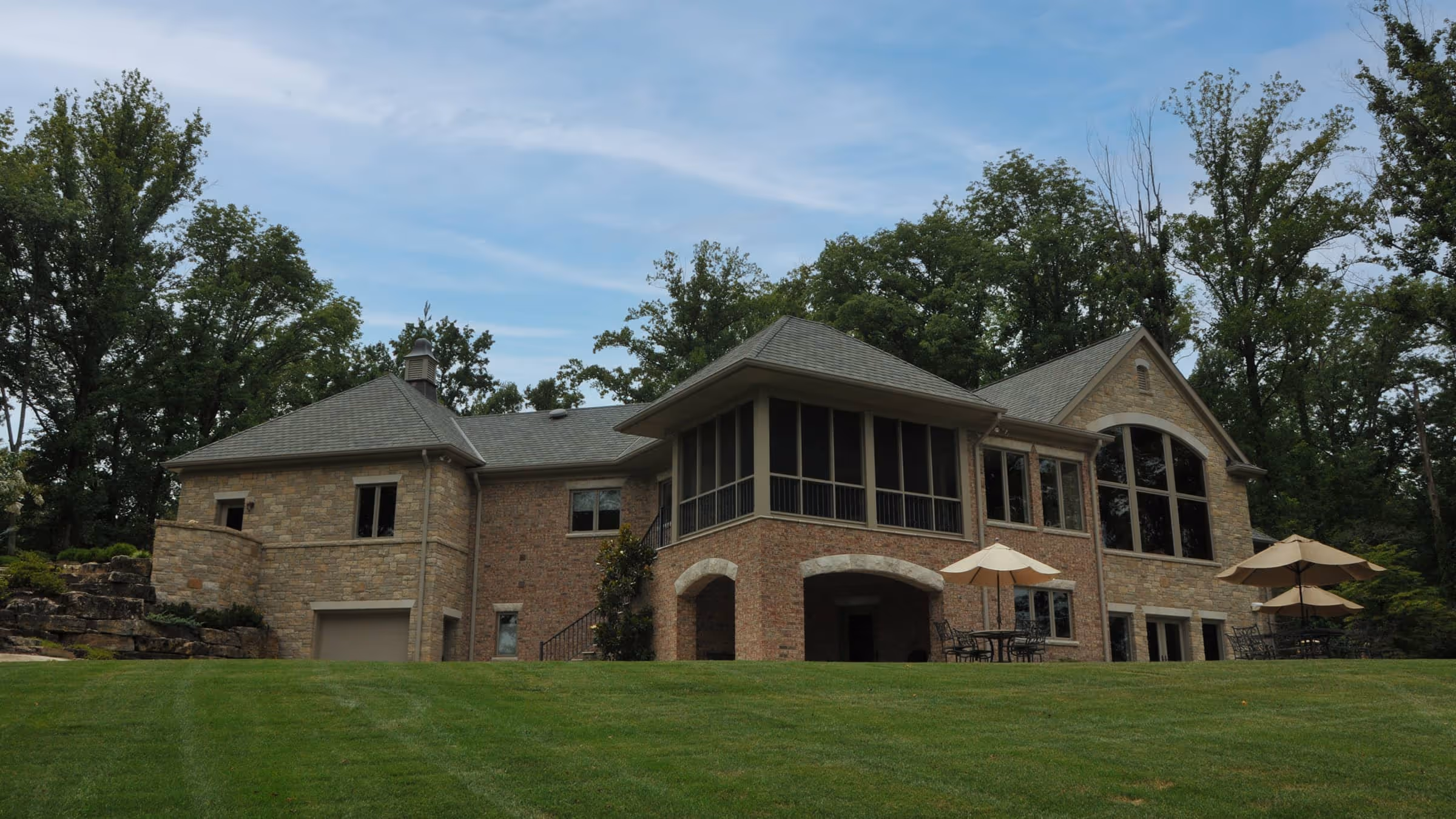 Large stone and brick house with a screened porch, patio tables with umbrellas, surrounded by green lawn and tall trees under a partly cloudy sky.