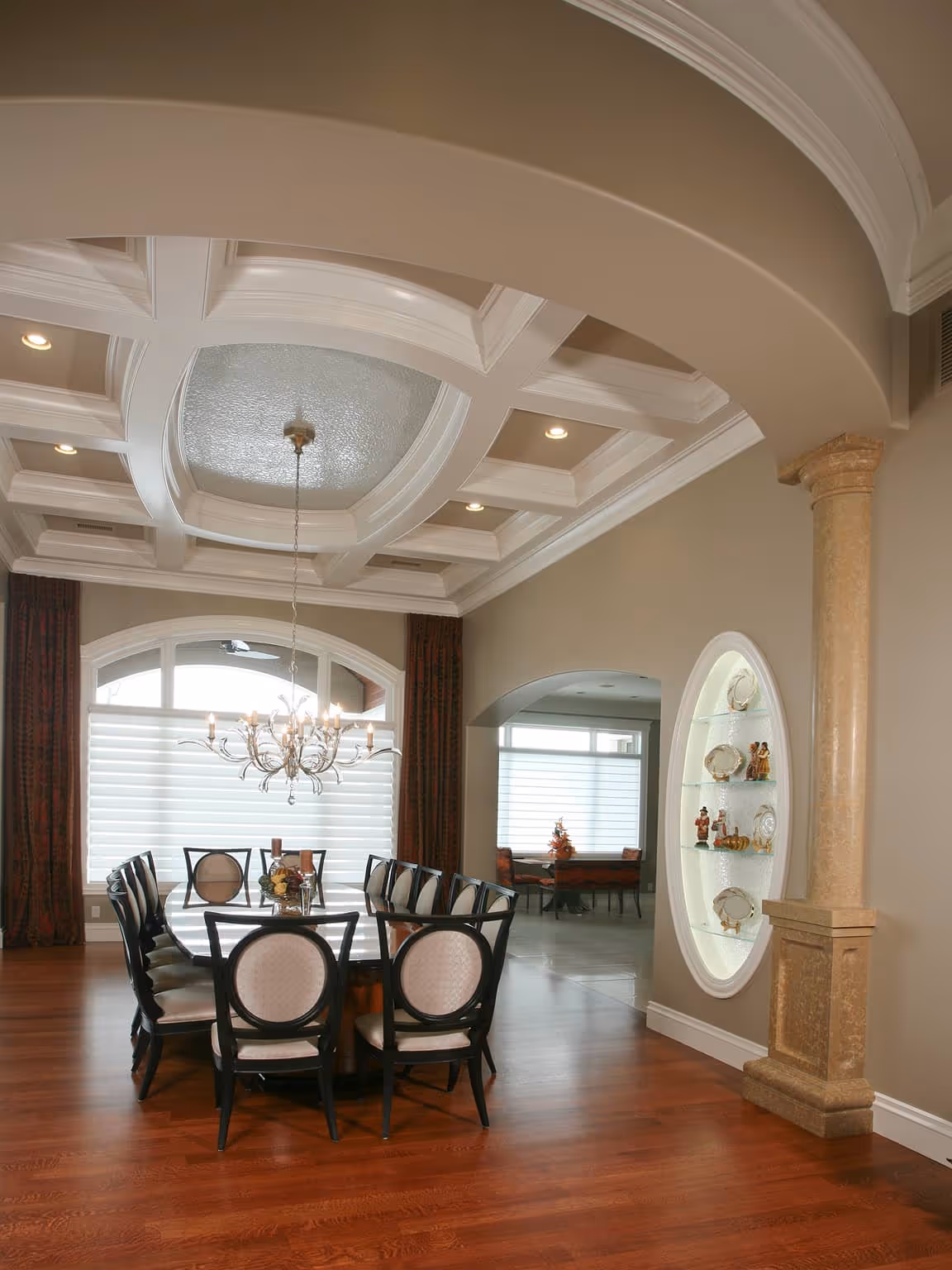 Elegant dining room with a long dark wood table surrounded by upholstered chairs, a decorative chandelier, coffered ceiling, and a built-in oval display shelf with figurines and plates.