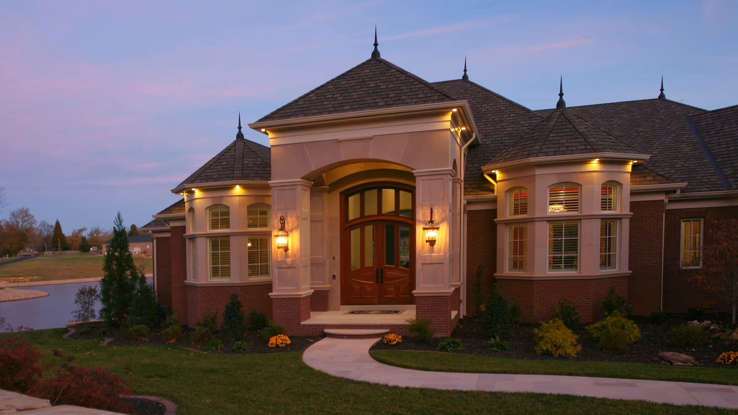 Elegant European Traditional style  house with warm exterior lights, a curved walkway, and landscaped front yard at dusk.