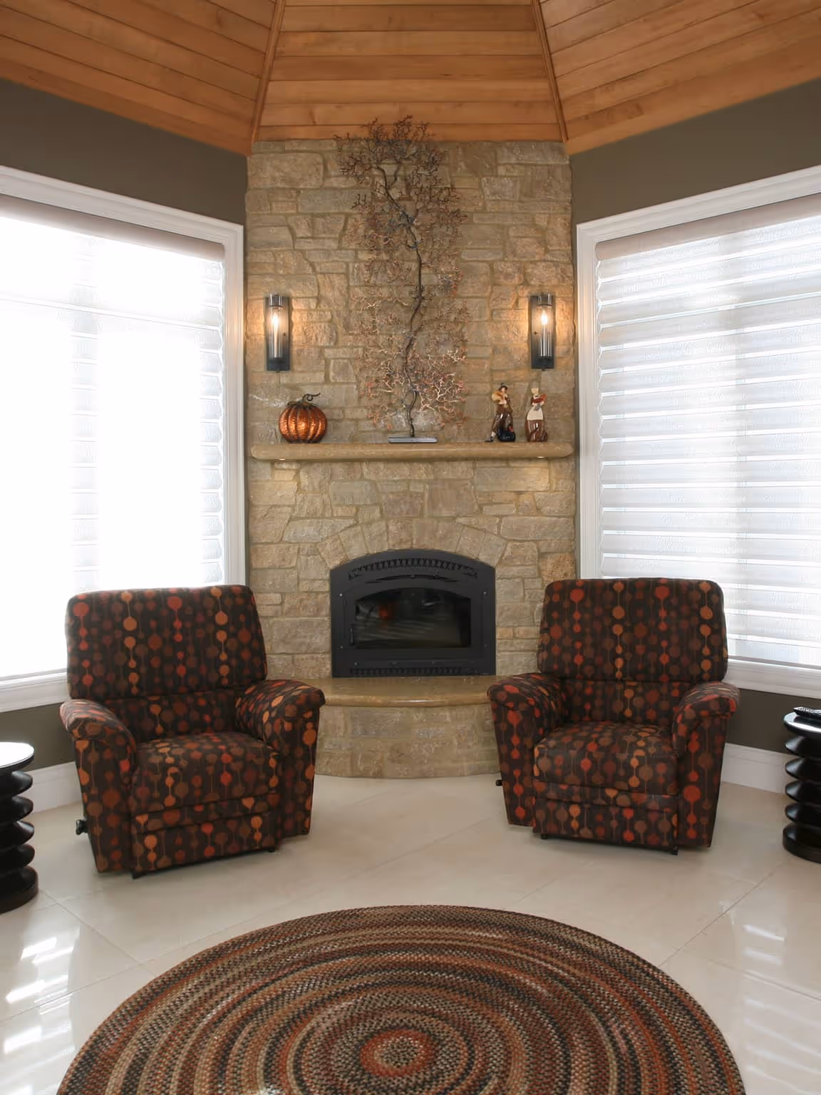 Cozy living room with stone fireplace, two patterned armchairs, wall sconces, and round braided rug.