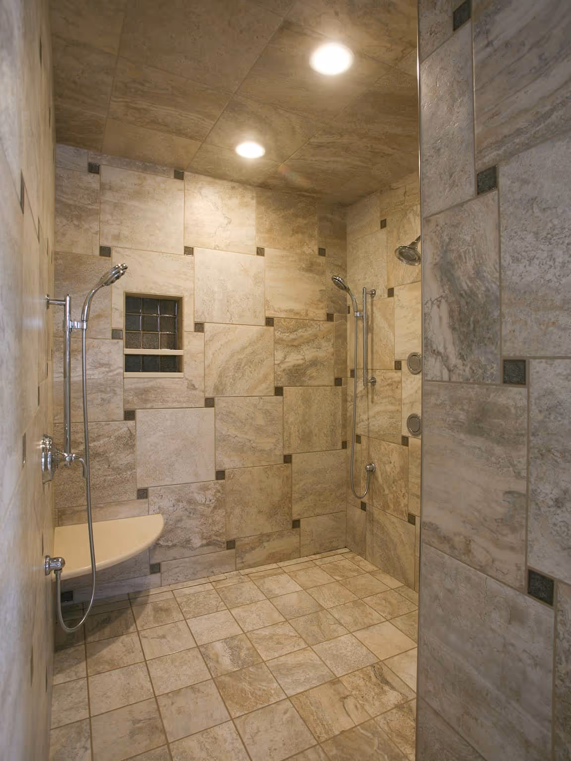 Spacious walk-in shower with beige stone tiles, two showerheads, and a small built-in bench.