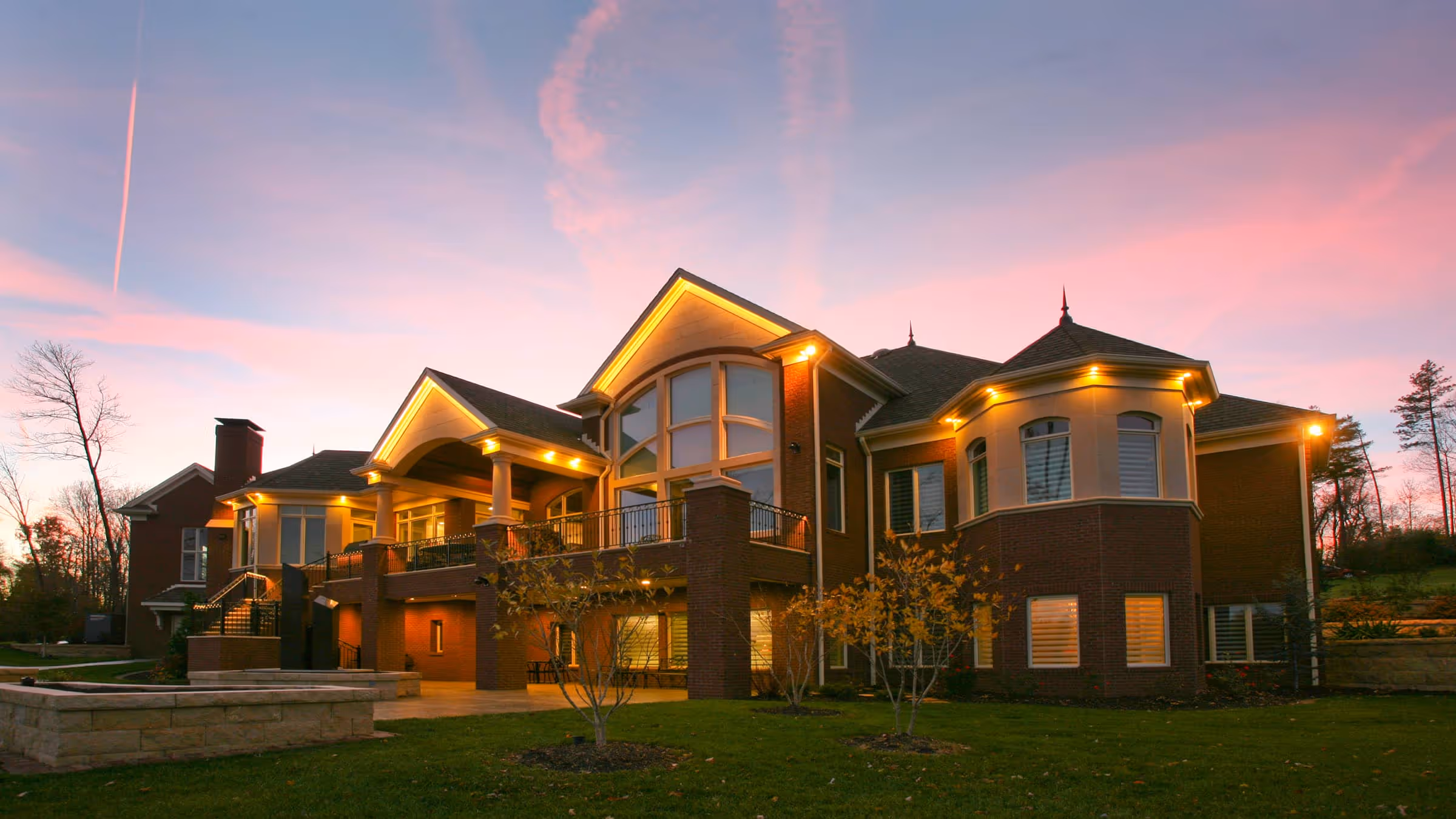 Large two-story European Traditional style  house illuminated with warm lights under a pink and purple sunset sky.