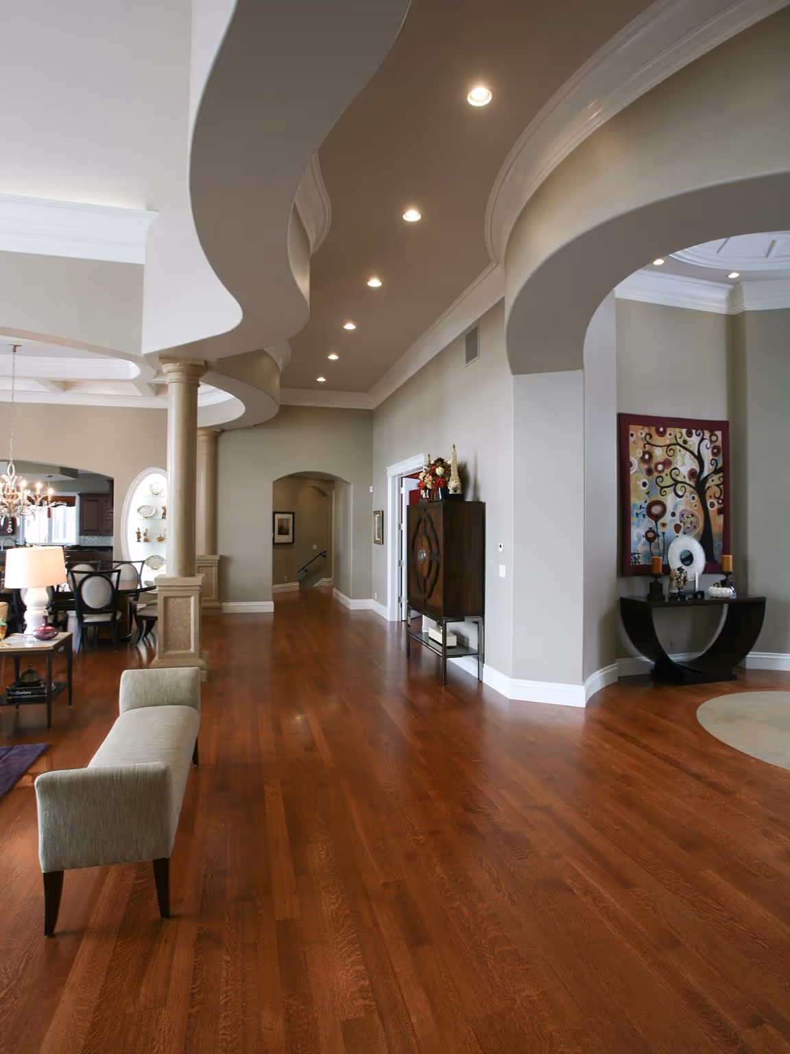 Spacious room with polished wooden floor, curved ceiling details, columns, a beige bench, and decorative furniture against light gray walls.