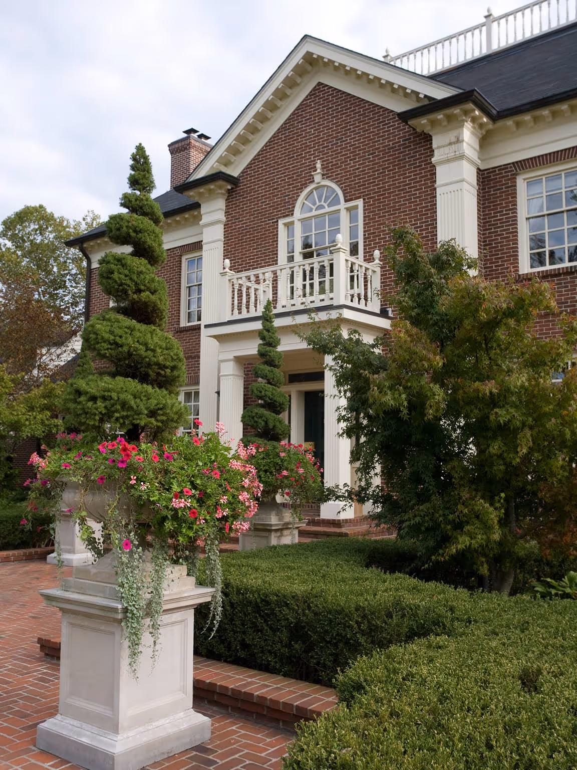 Georgian style brick house with white columns, a small balcony, manicured bushes, and topiary trees in stone planters with pink flowers.