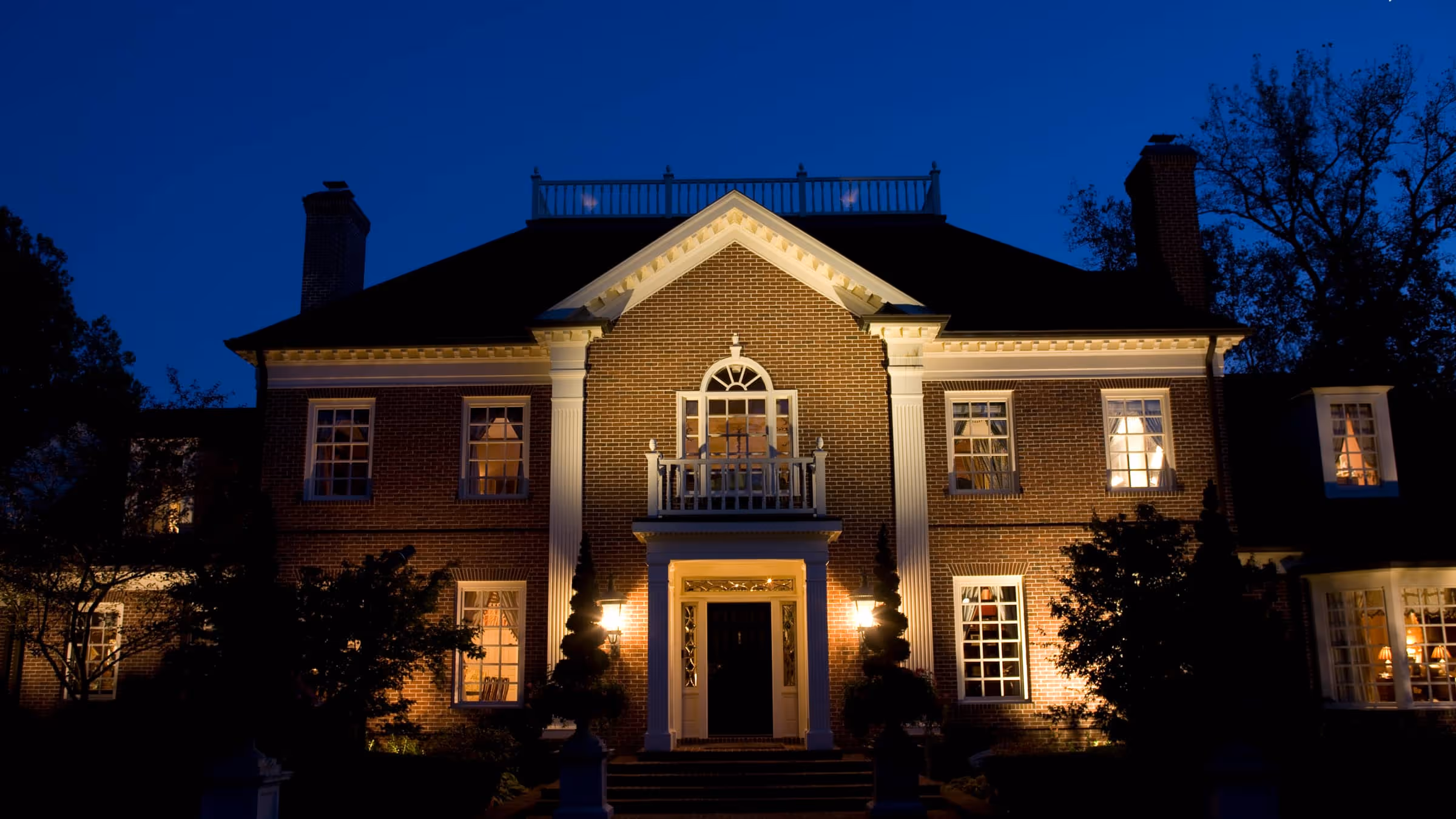Illuminated Georgian style brick house at night with white columns and multiple windows showing interior lights.