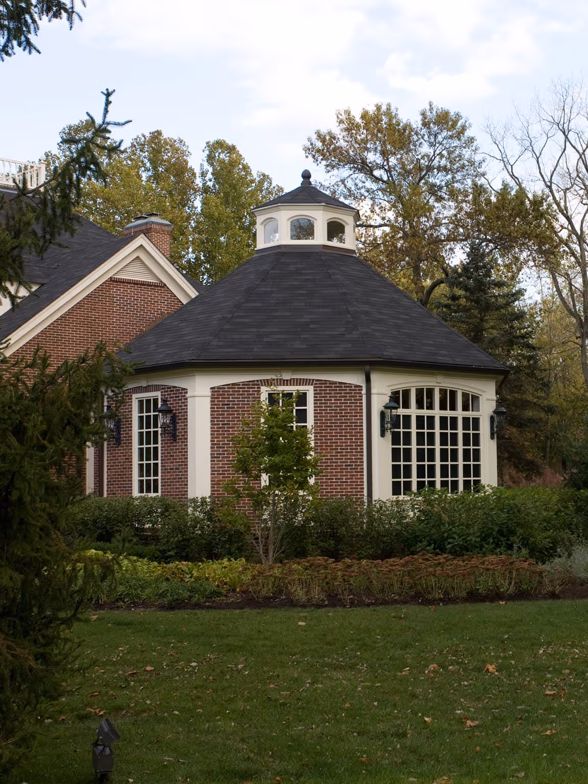 Brick Georgian style gazebo with white trim and a dark shingled roof surrounded by green shrubs and trees.
