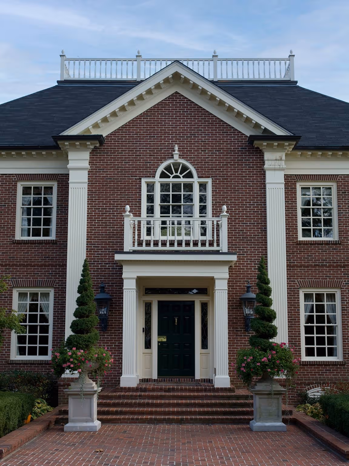 Front entrance of a Georgian style brick house with white columns, arched window above the doorway, and spiral topiary plants in planters.