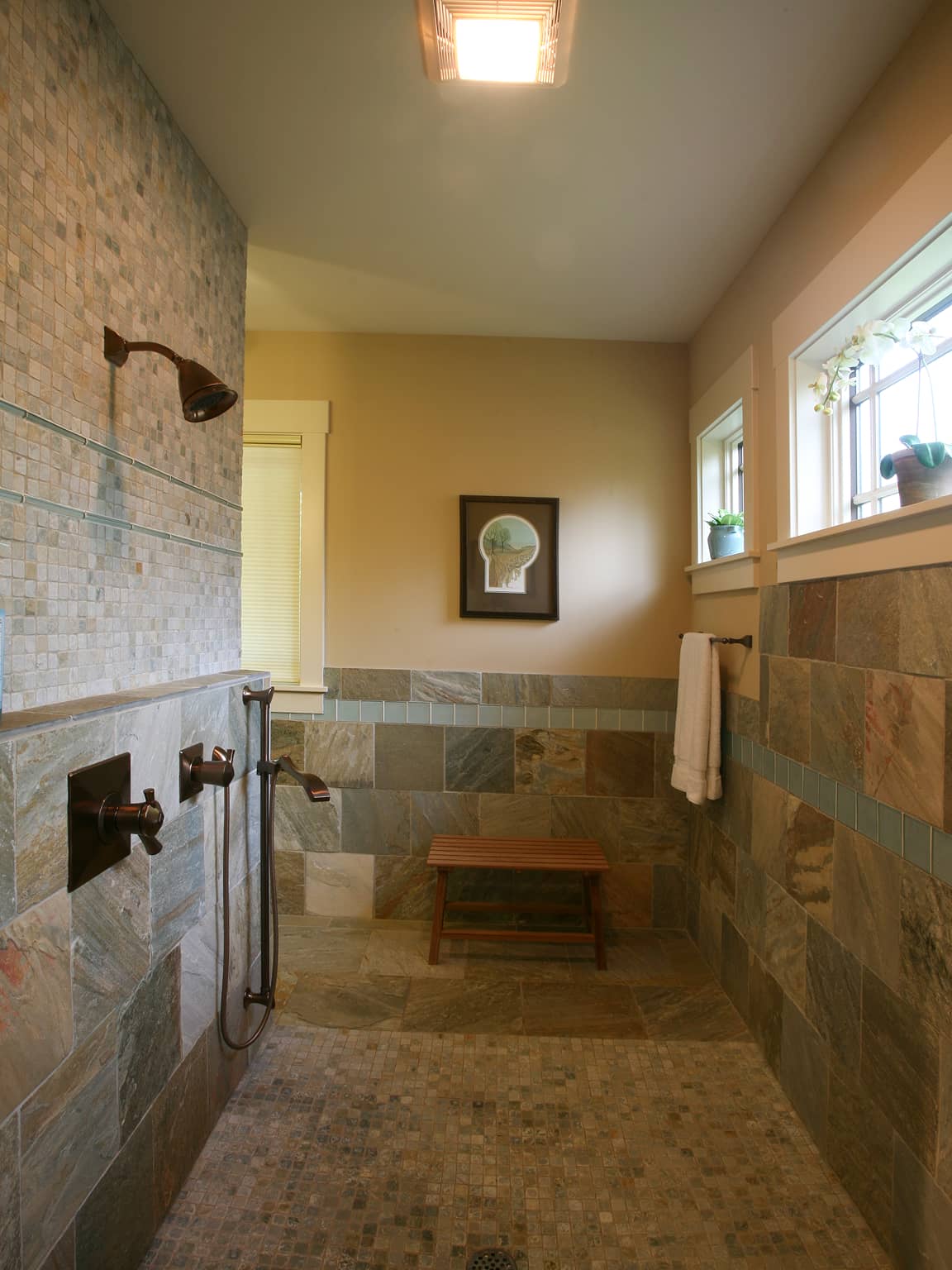 Walk-in shower with stone tiles, bronze fixtures, a wooden bench, and windows with small potted plants.