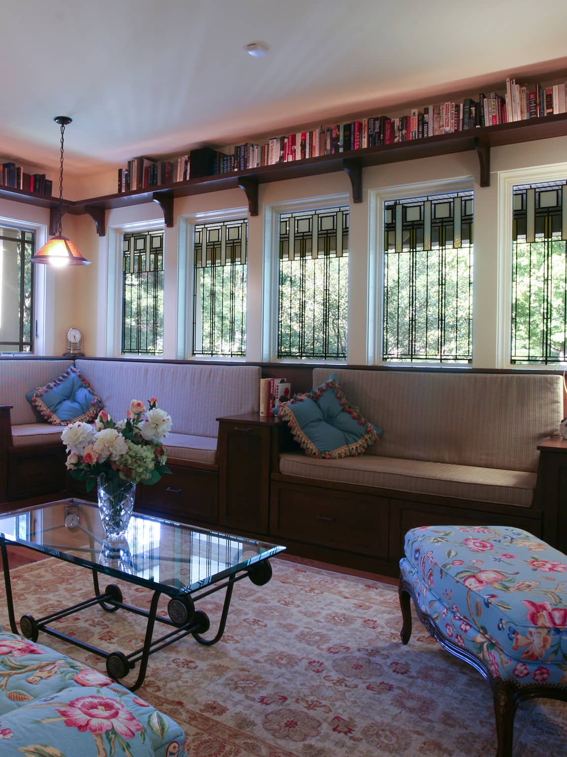 Cozy living room with stained glass windows, built-in bench seating with cushions, floral ottoman, glass coffee table with flower vase, and bookshelf above windows.