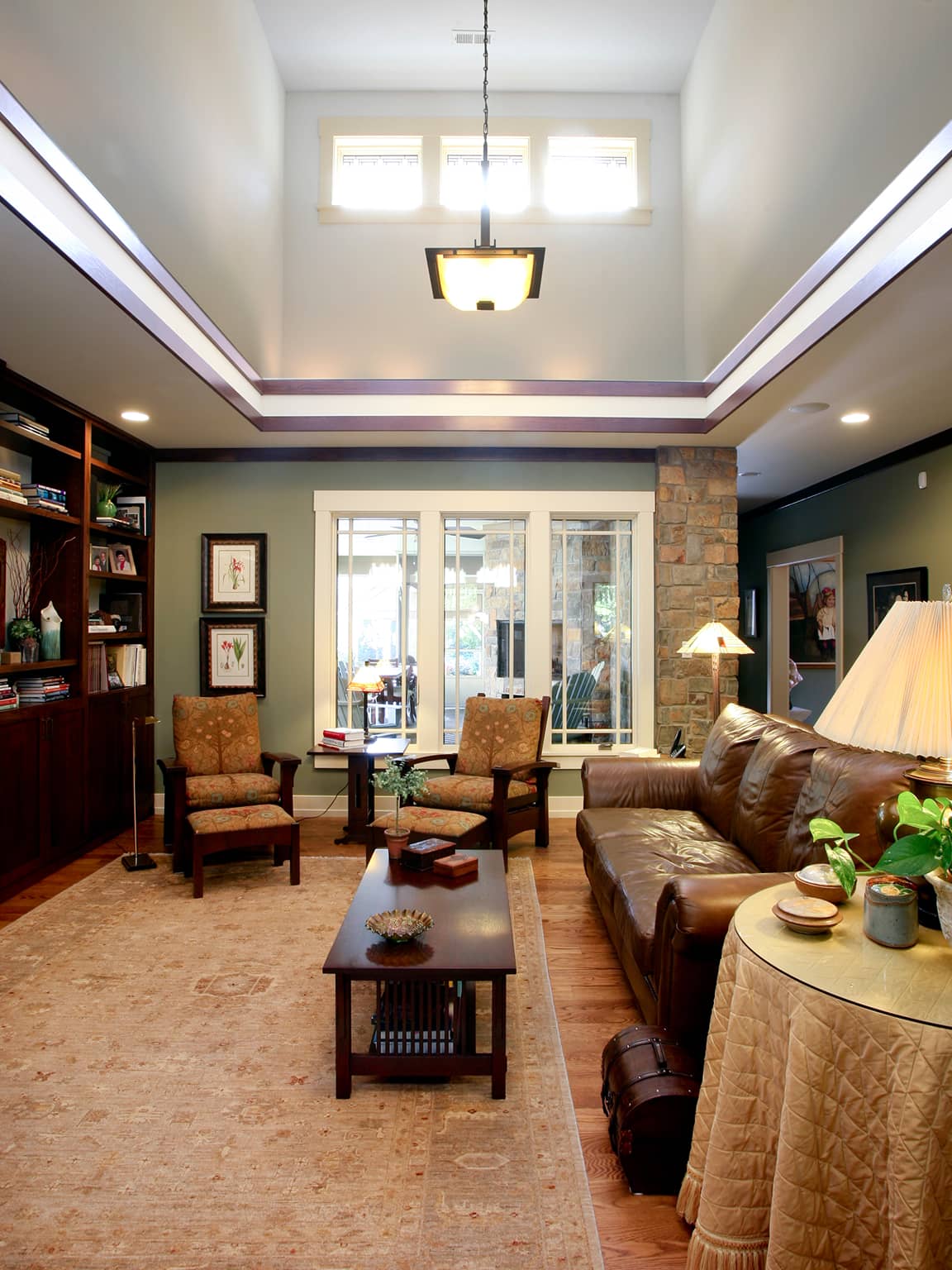 Cozy living room with high ceilings, brown leather sofa, two patterned armchairs, wooden coffee table, large window, and built-in bookshelf.