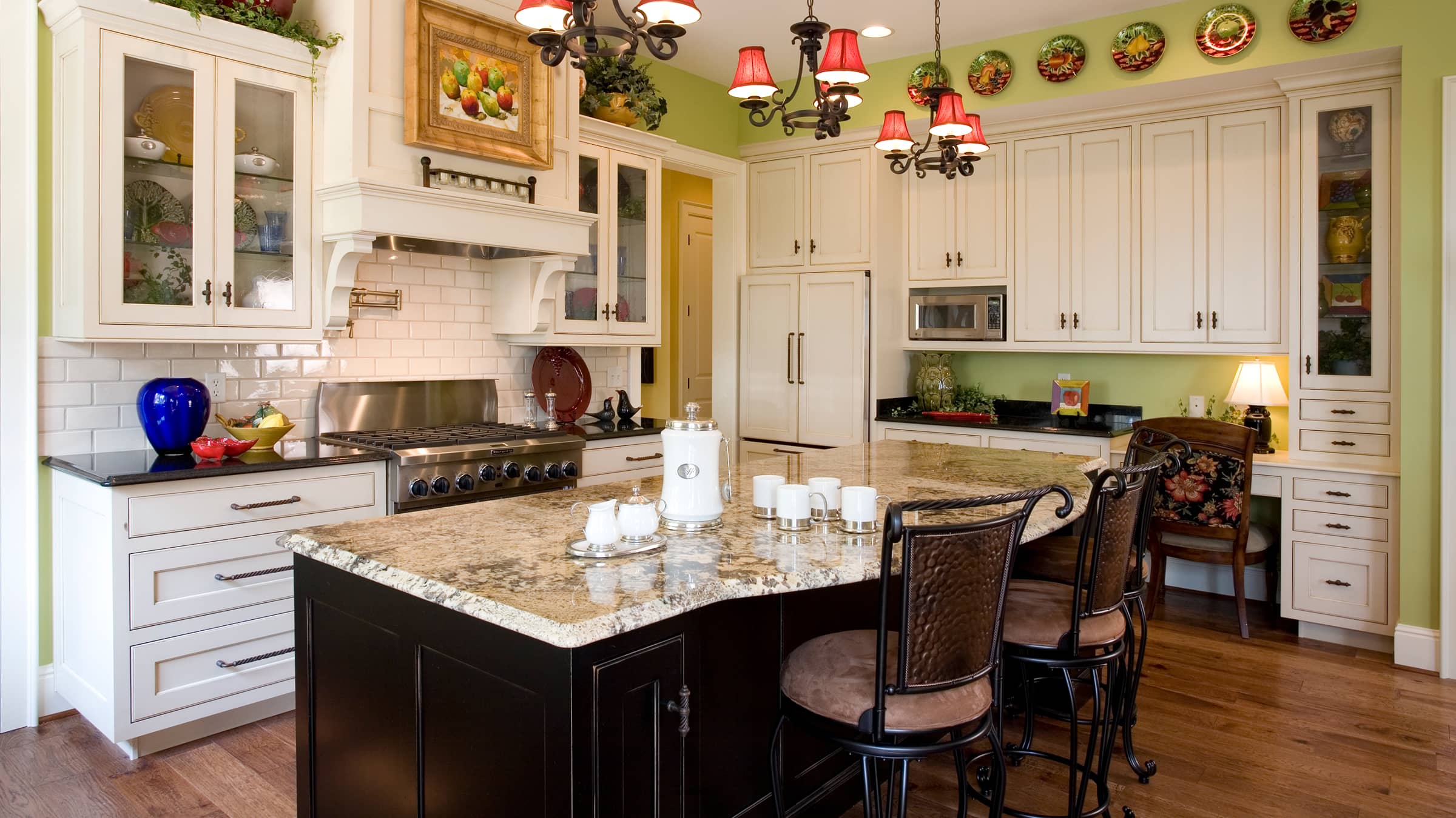 Spacious kitchen with a large granite island, black base cabinets, white upper cabinets, stove with stainless steel hood, and decorative plates on a green wall.
