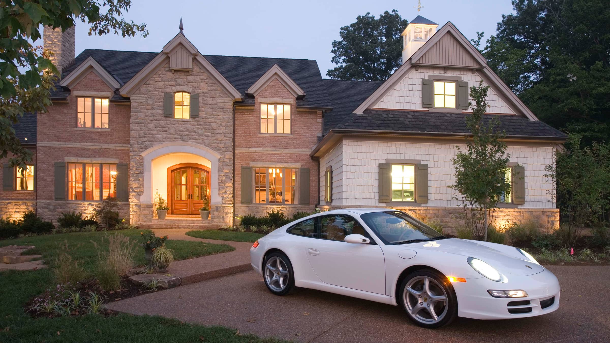 White Porsche sports car parked on driveway in front of a large, lit, two-story European Country Estate style house at dusk.