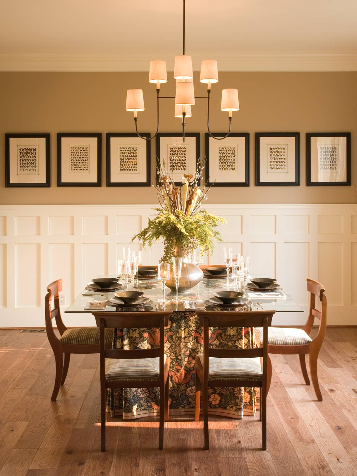 Elegant dining room with glass table, floral tablecloth, six wooden chairs, black tableware, a large floral centerpiece, and framed butterfly prints on the beige wall.