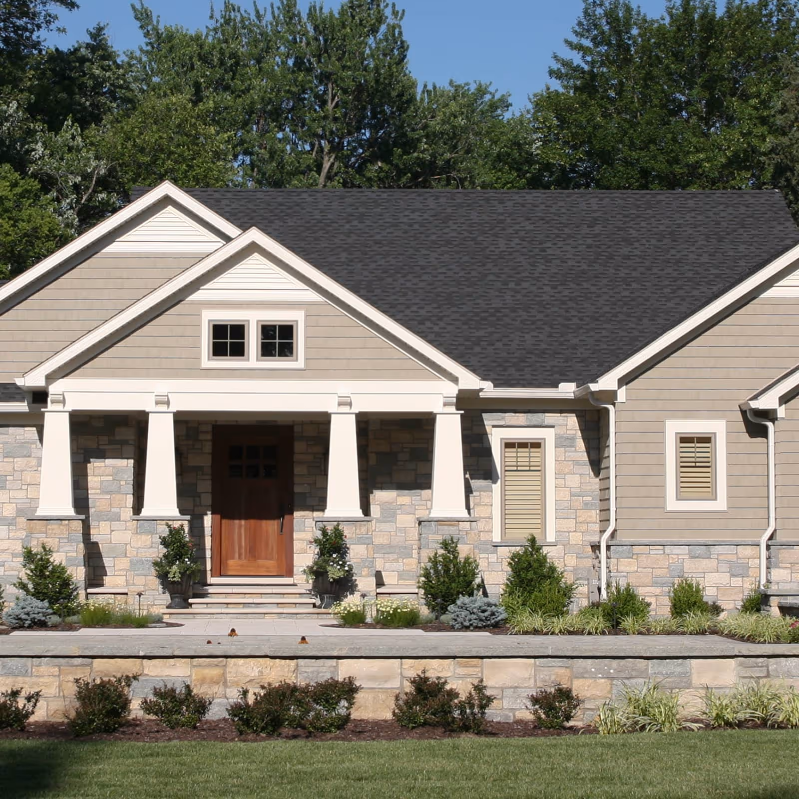 Craftsman-style house front with stone and beige siding, tapered white columns, dark roof, and landscaped yard.
