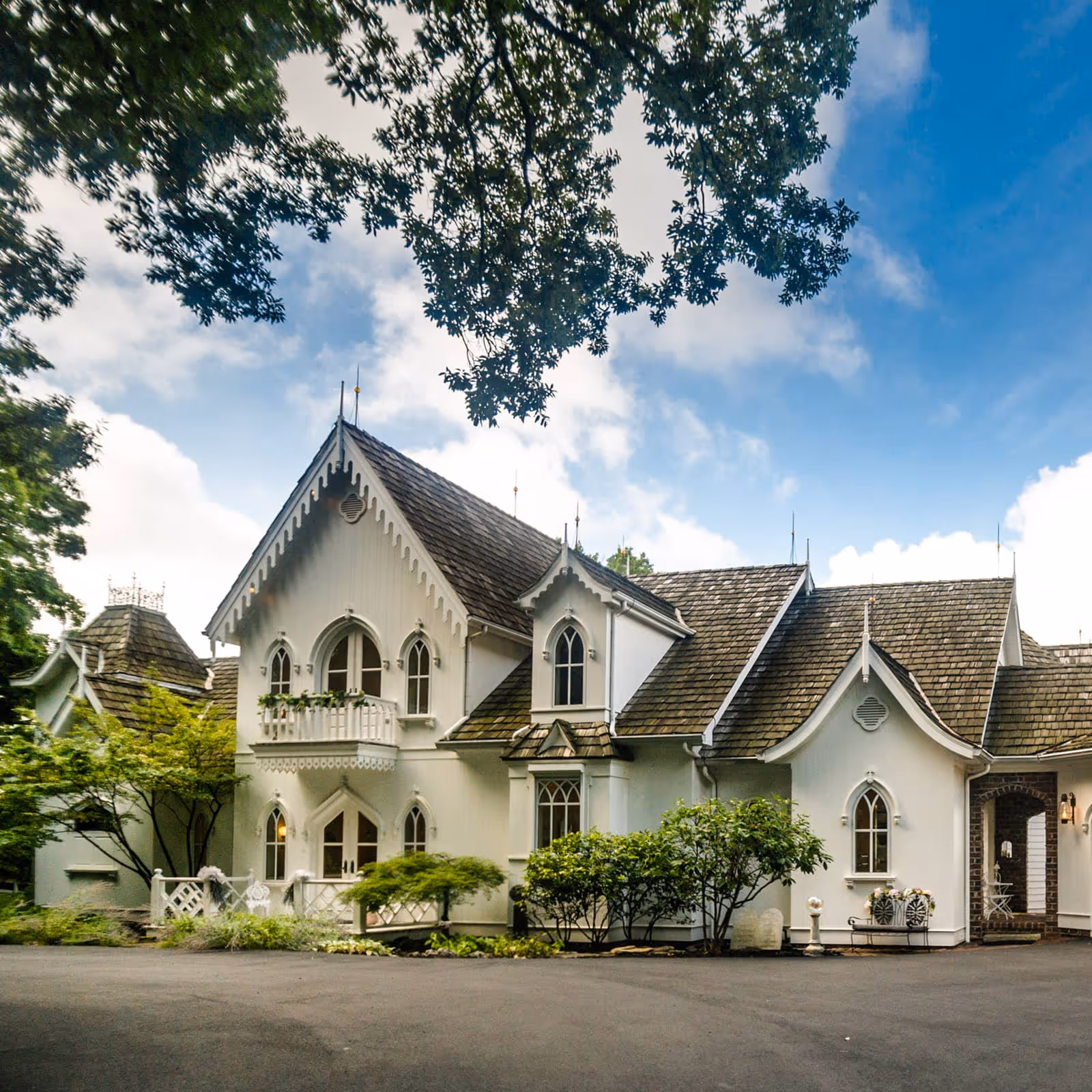 White Victorian-style house with steep gabled roofs and arched windows surrounded by trees under a partly cloudy blue sky.