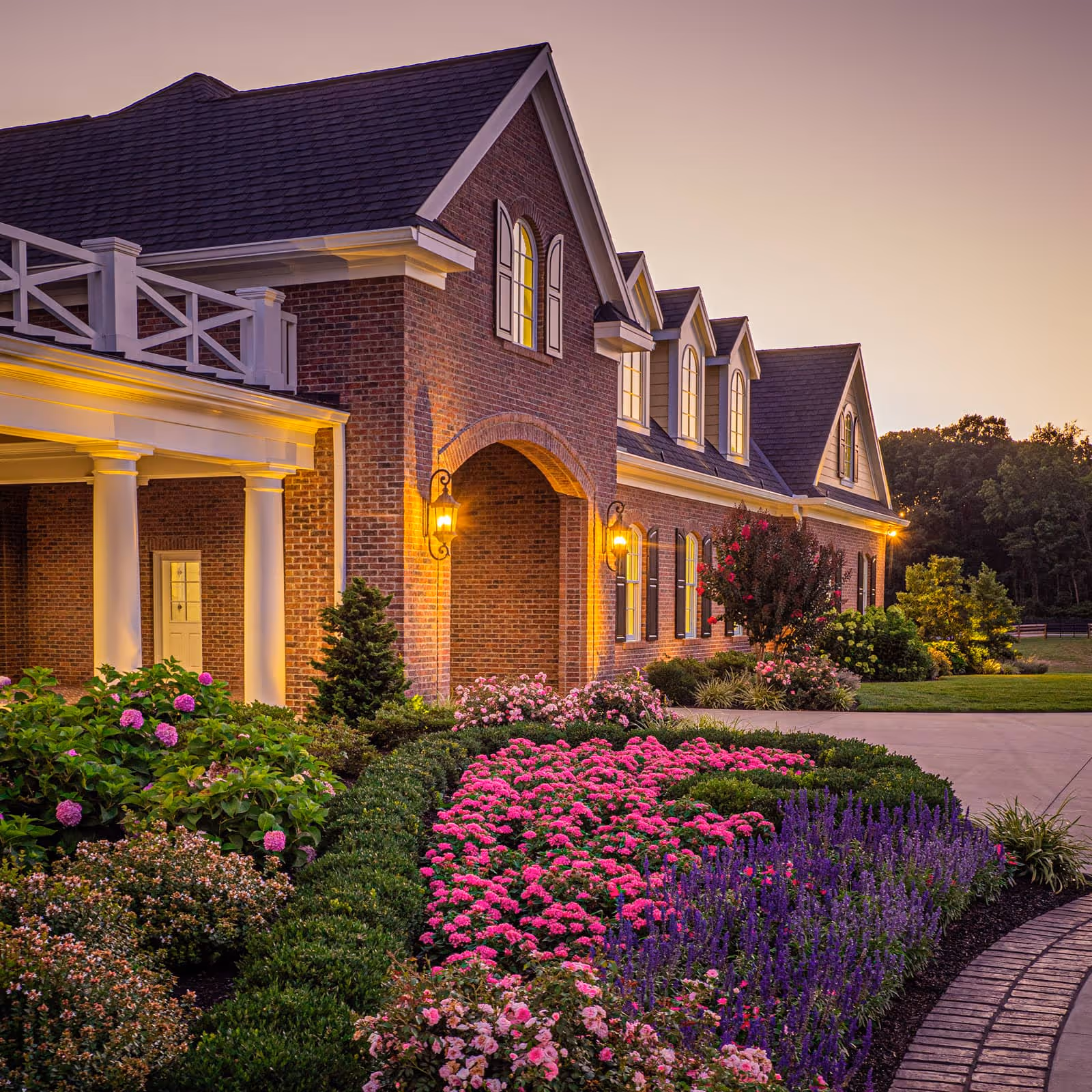 Large brick house with lit windows and porch lights, surrounded by colorful flower beds and manicured shrubs during sunset.