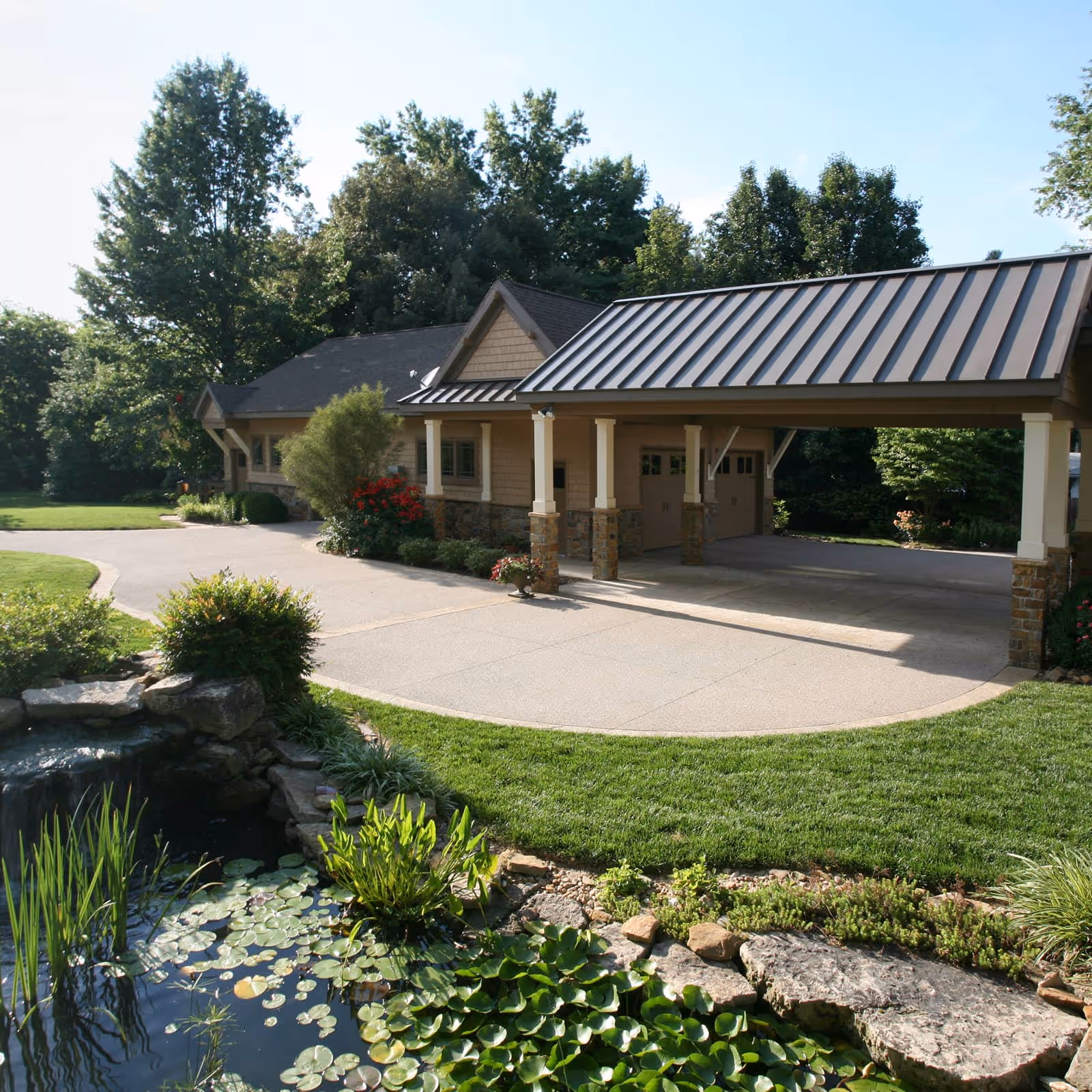 House with a covered driveway, surrounded by green trees, a manicured lawn, and a pond with lily pads and rocks in the foreground.