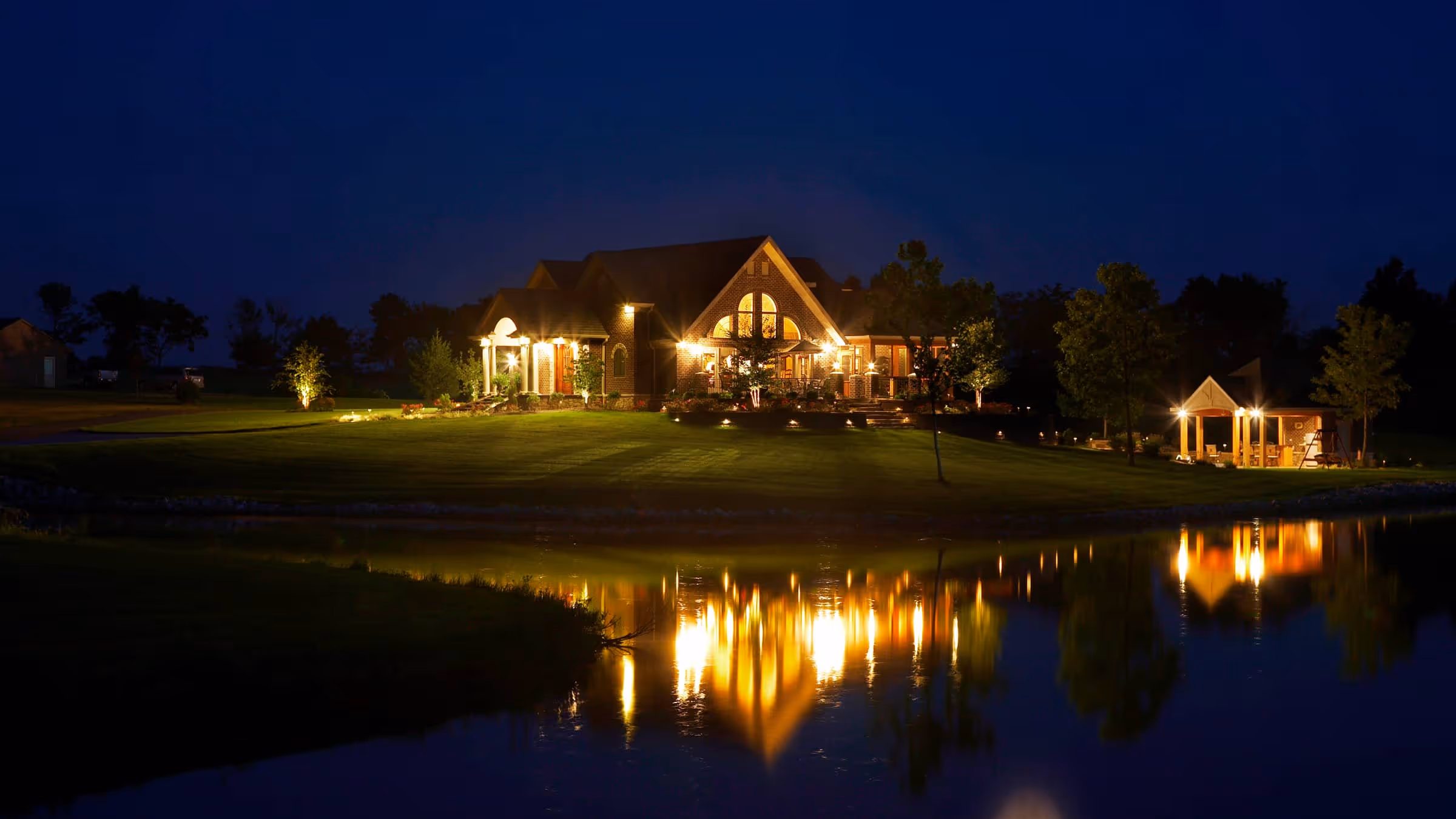 A large, well-lit brick Traditional style house at night with its lights reflecting on a nearby lake.