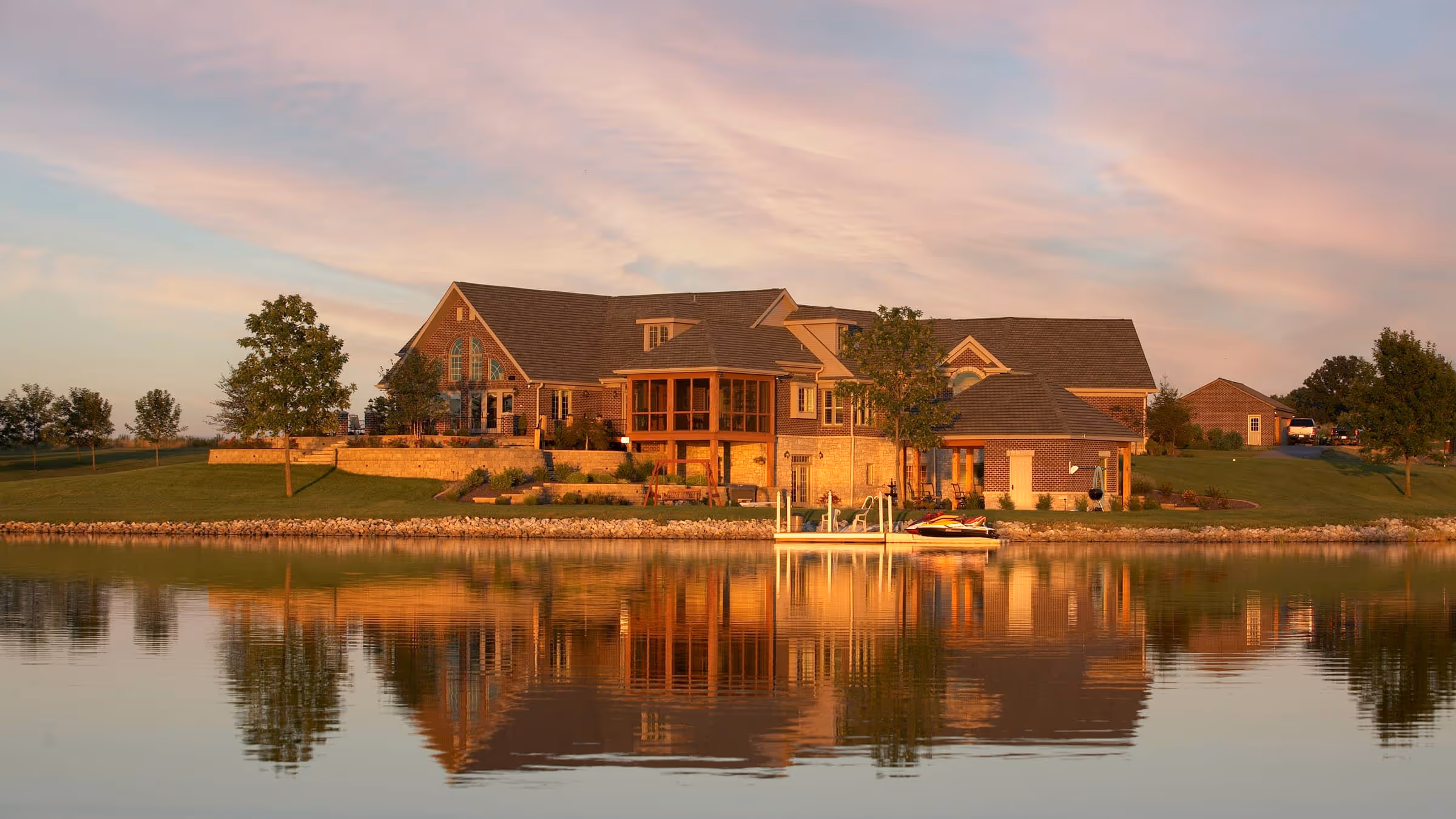 Large brick and stone Traditional style house on a grassy lakeshore reflected in calm water at sunset.