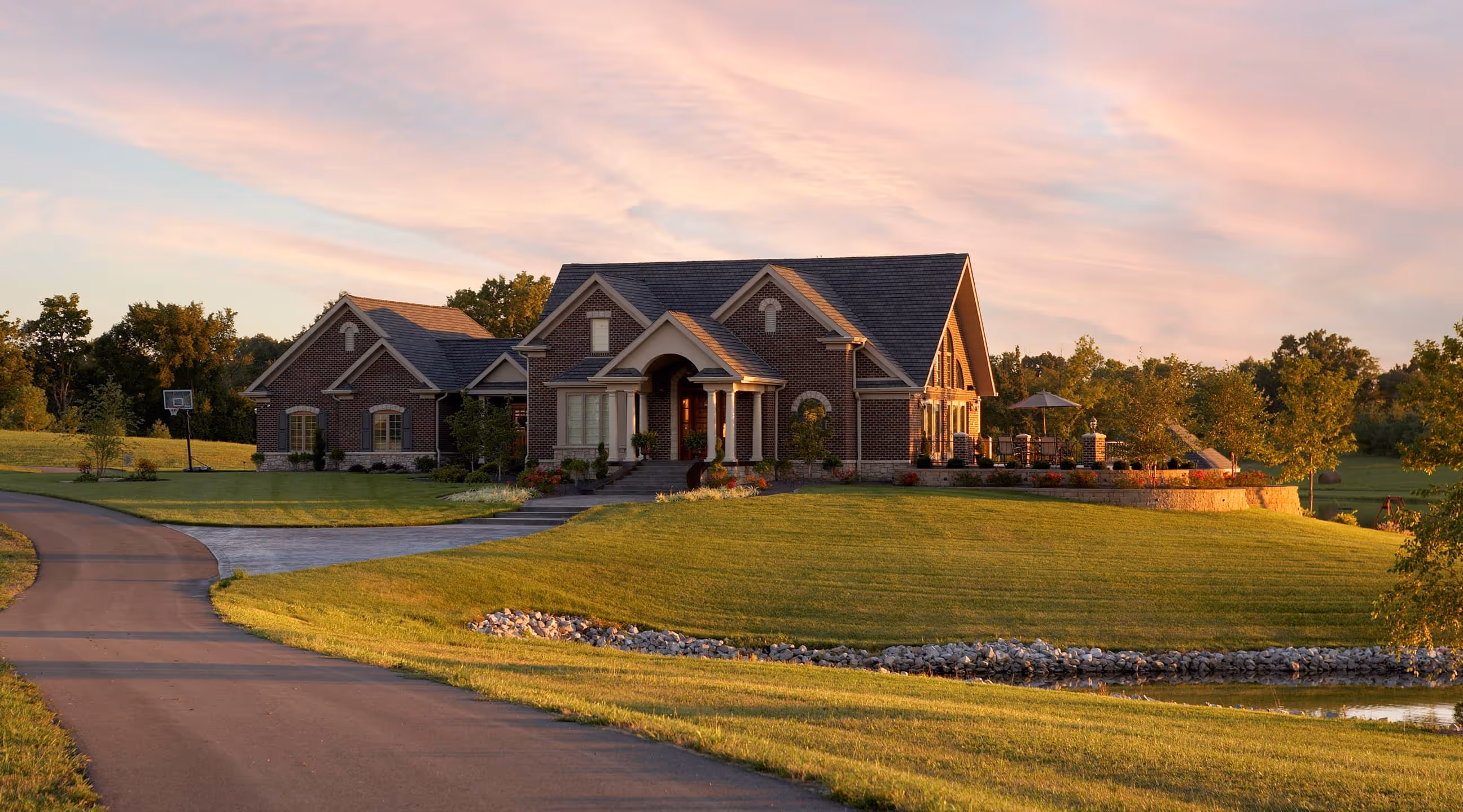 Large brick Traditional style house with a manicured lawn, driveway, and sunlit patio in a rural setting at sunset.