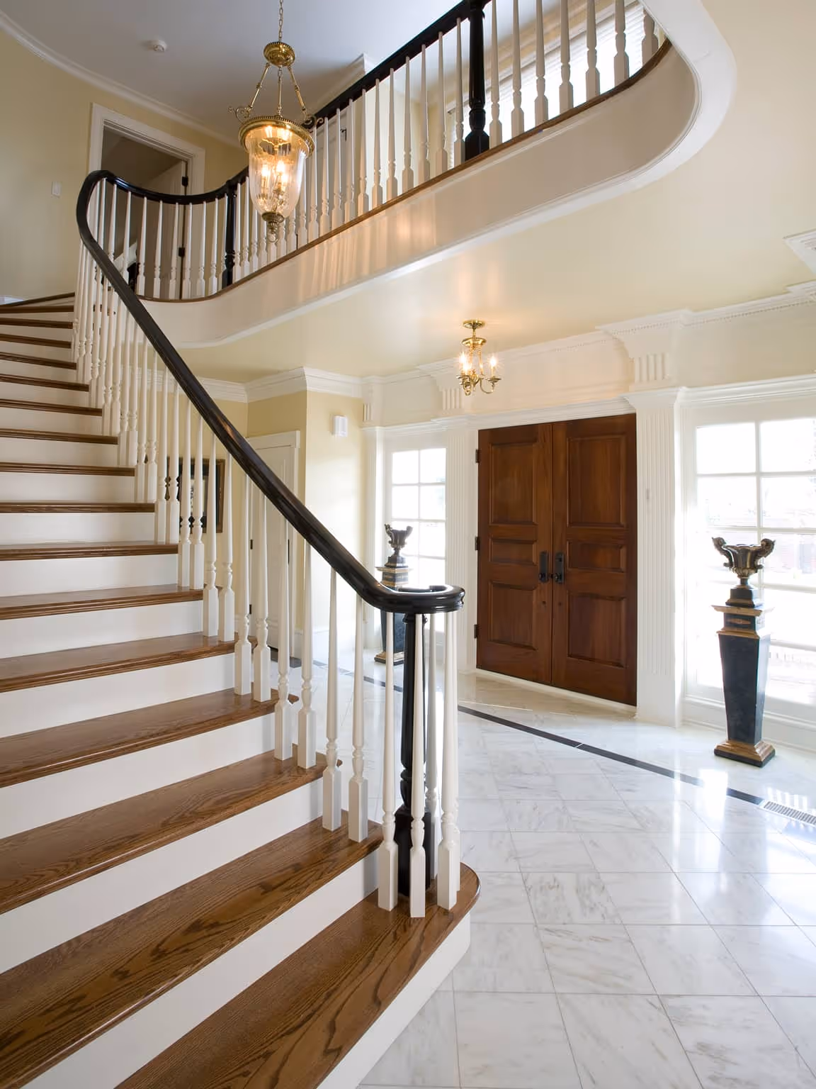 Elegant staircase with wooden steps and white railing in a spacious foyer with marble floor and double wooden doors.