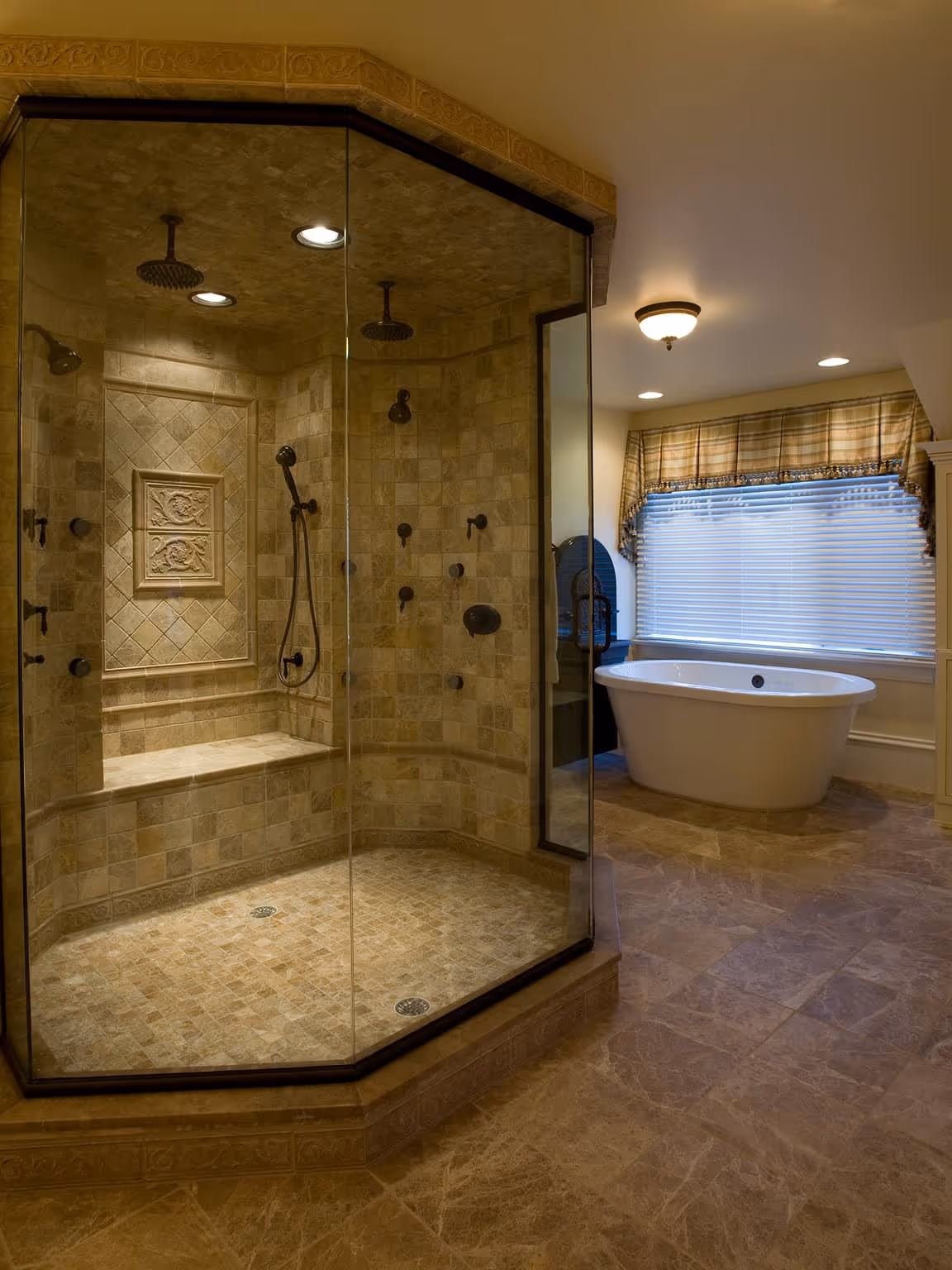 Spacious bathroom featuring a large glass-enclosed tiled shower with multiple showerheads and a built-in bench, and a freestanding white bathtub near a window with blinds and a plaid valance.