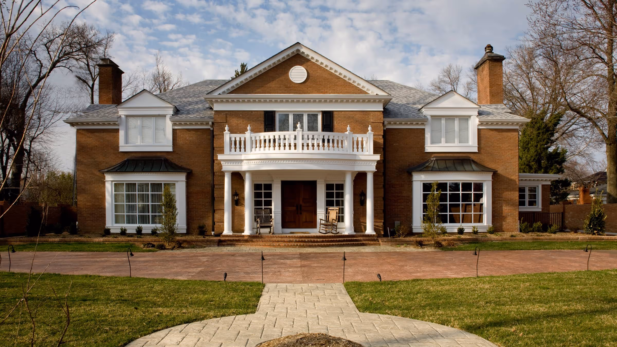 Large two-story brick Early Classical Revival style house with white columns, two chimneys, and a balcony above the front porch with rocking chairs.