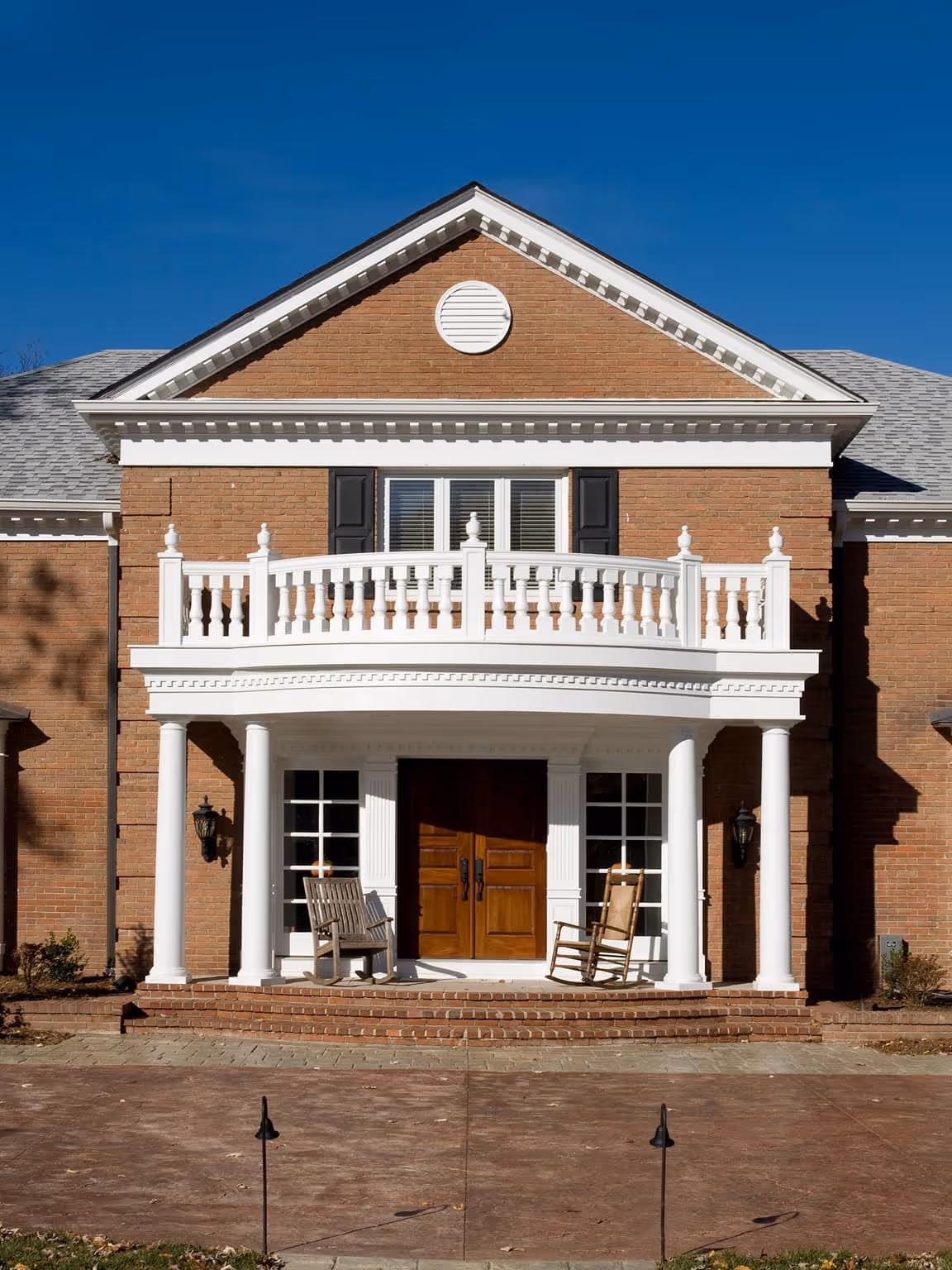 Brick Early Classical Revival style house with white classical columns, balcony railing, and wooden double front doors with two chairs on the porch.