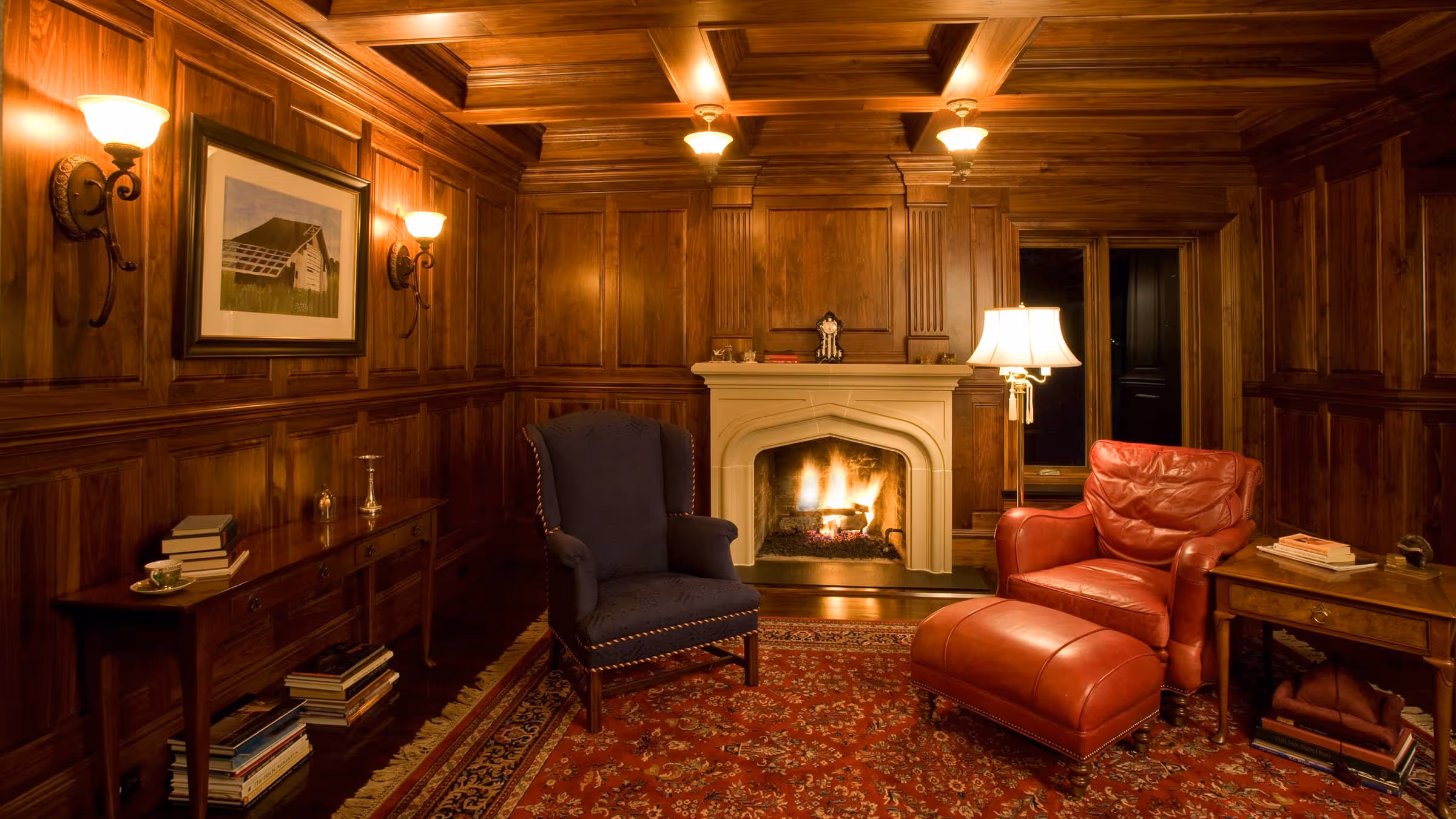 Cozy room with wooden panel walls, a lit fireplace, a blue upholstered armchair, a red leather armchair with matching ottoman, and books on side tables.