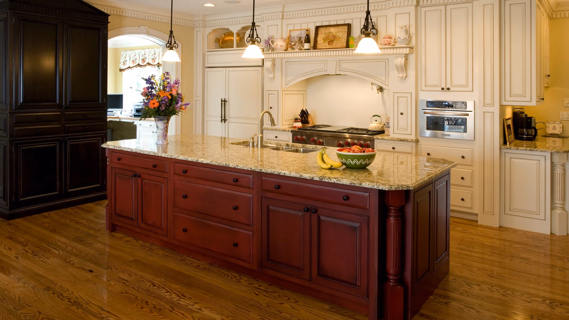 Spacious kitchen with a large granite countertop island featuring a flower vase, bananas, and a bowl of apples, surrounded by cream and dark wood cabinetry.