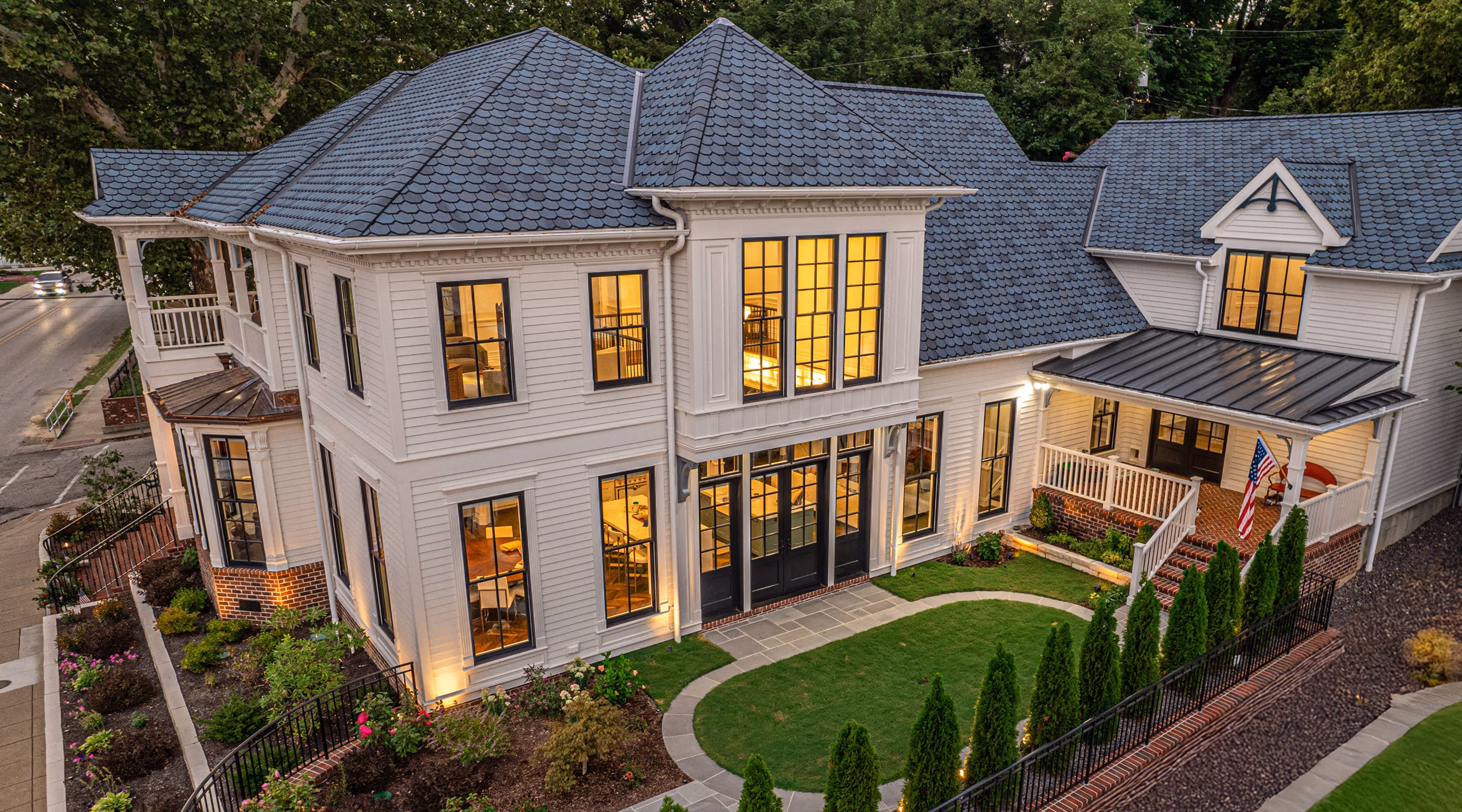 Modern Victorian style two-story house with white siding, black framed windows, blue-gray shingle roof, and lit interior at dusk surrounded by landscaped garden and greenery.