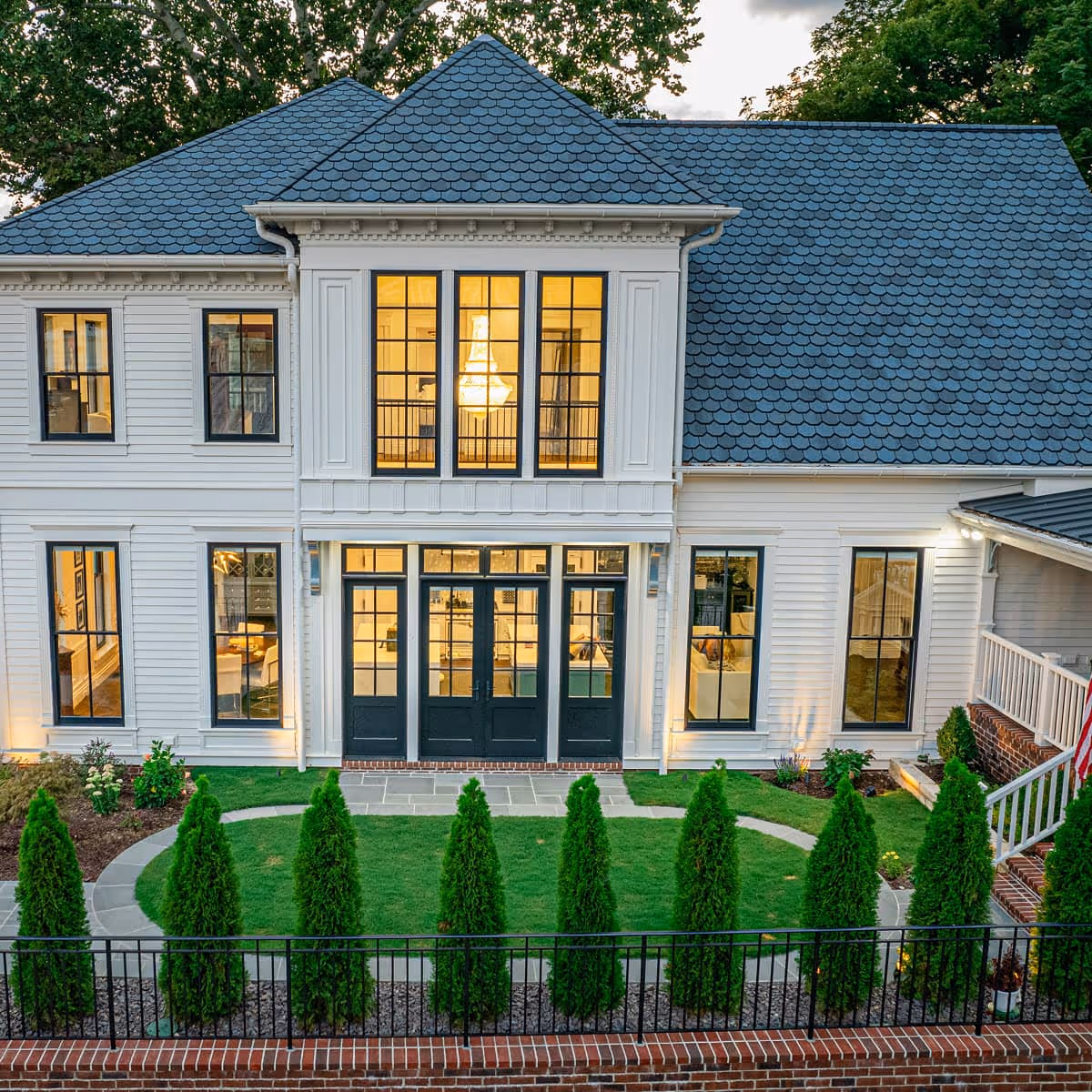 Modern Victorian white house with large windows, blue scalloped roof, front lawn with tall shrubs, and black front doors with glass panels.