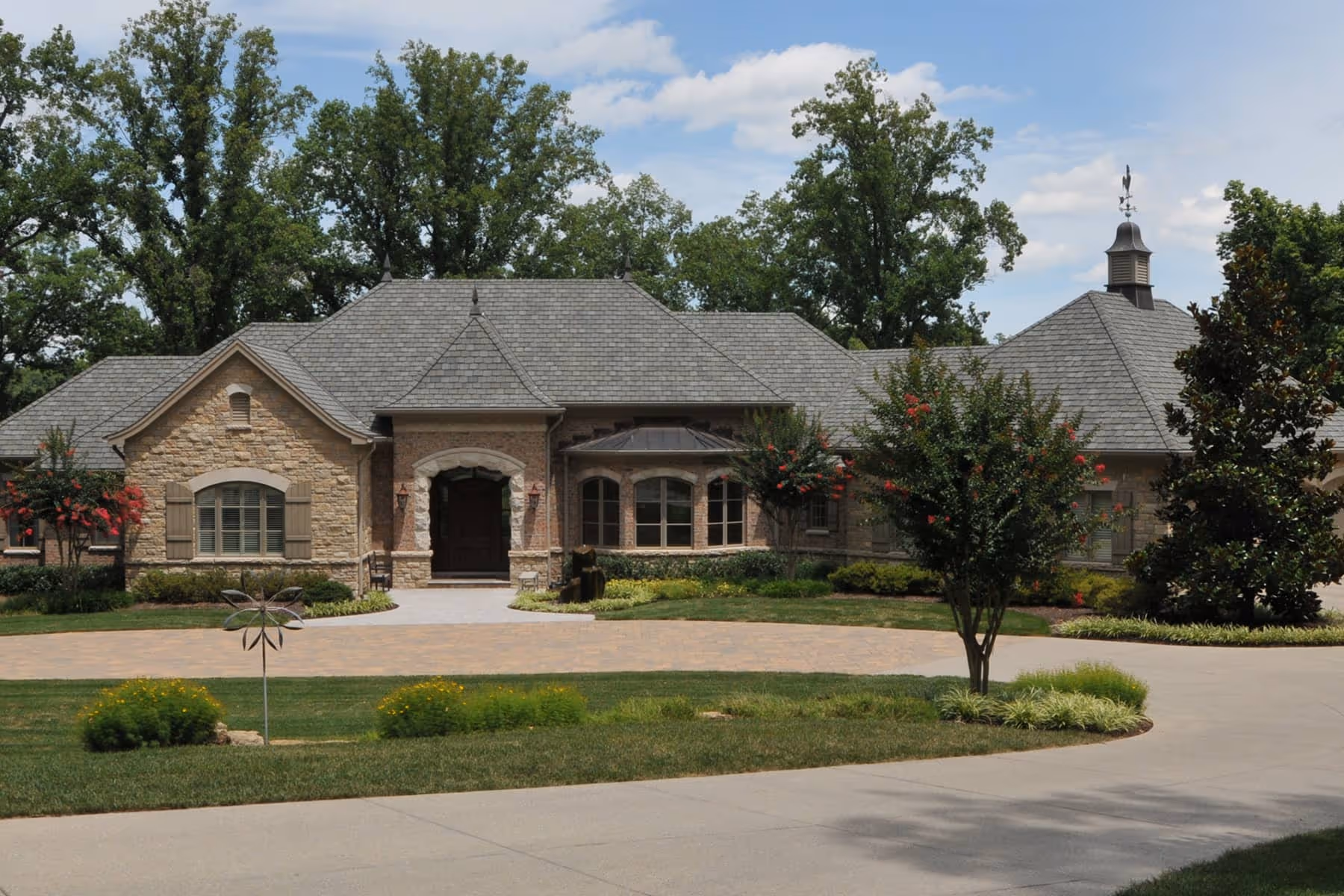 Large stone and brick country French style house with gray shingled roof and landscaped front yard with trees and shrubs.