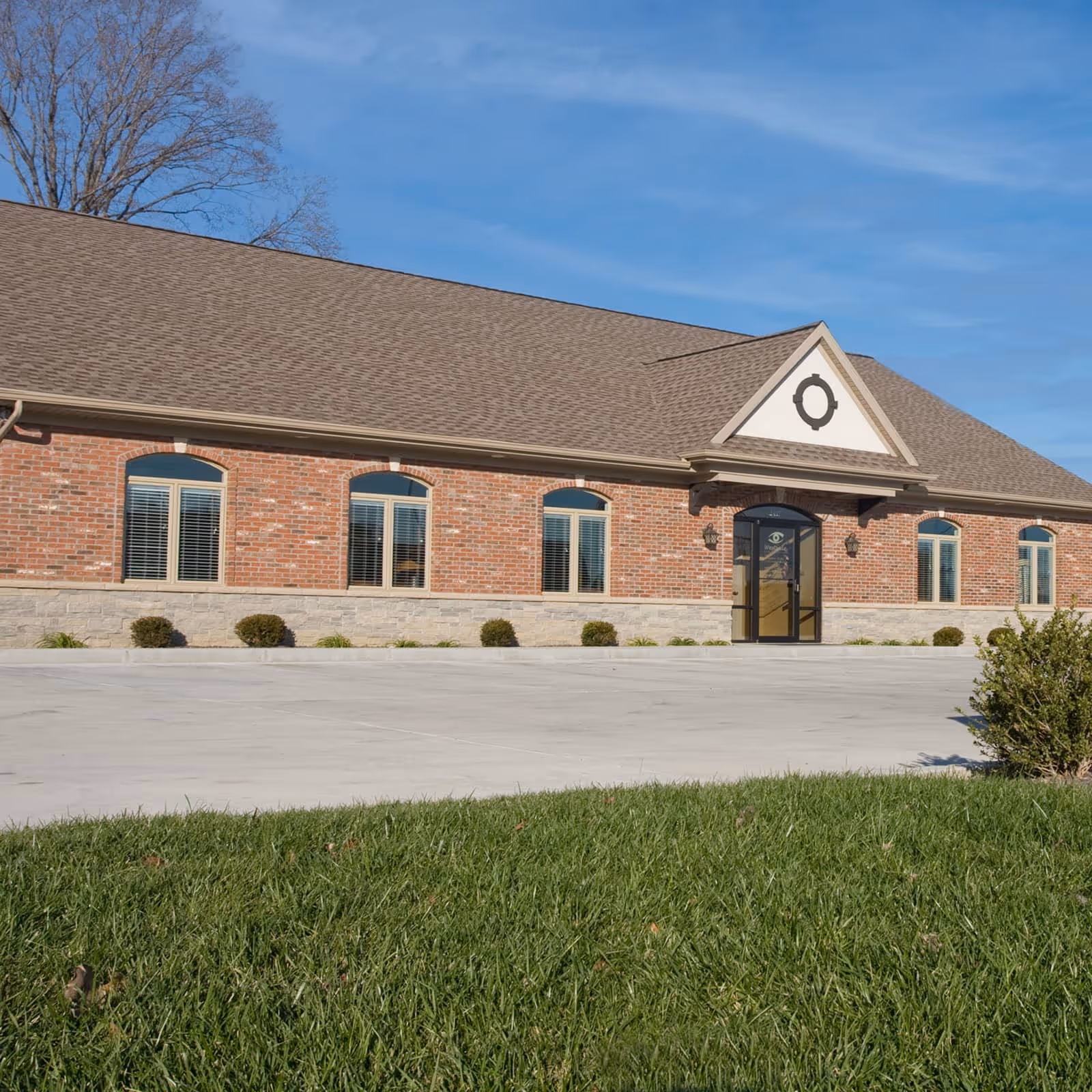 Single-story brick office building with large windows, central glass door, surrounded by low bushes and a green lawn under a blue sky.
