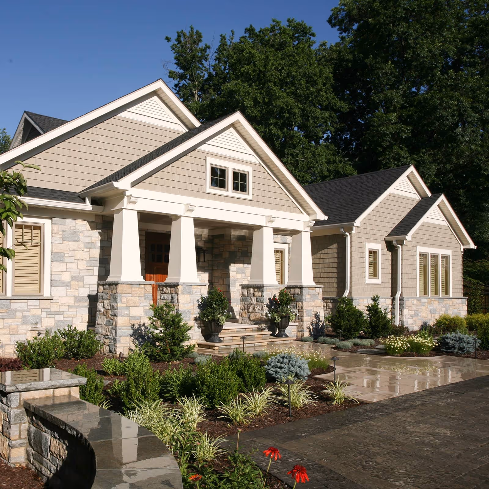 Craftsman-style house with stone pillars, beige siding, landscaped front yard, and clear blue sky.