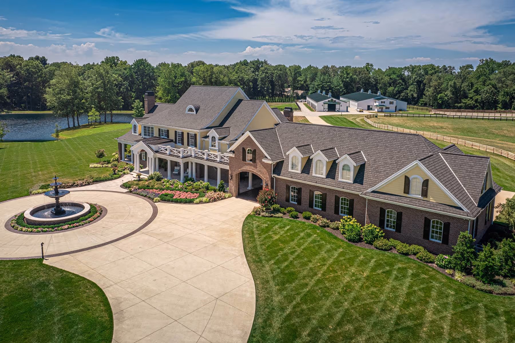 Large southern-style estate with brick and yellow siding, a circular driveway with central fountain, manicured lawns, flower beds, and a pond with wooded background.