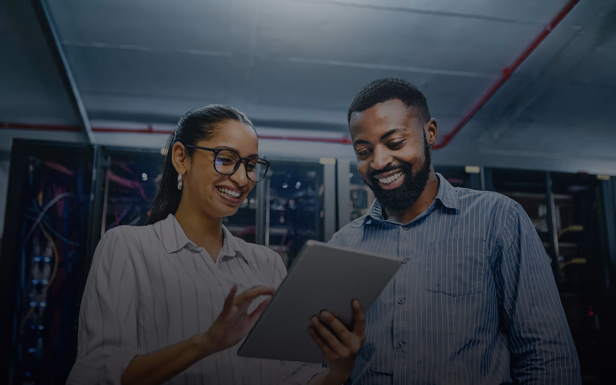 Two smiling professionals, a woman and a man, looking at a tablet in a server room.