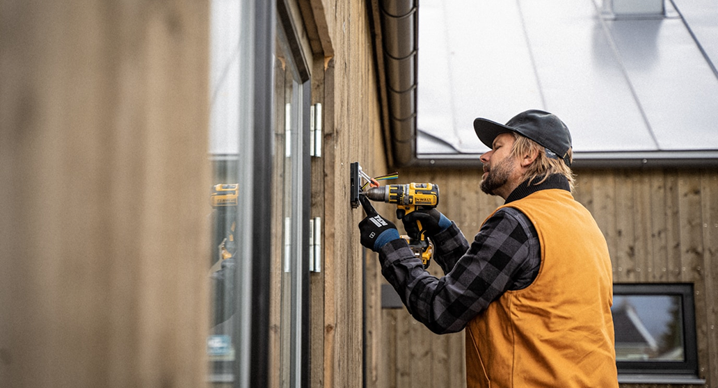 Man wearing a cap and orange vest using a cordless drill to install or fix a device on wooden exterior wall.