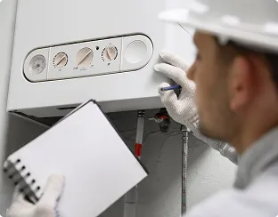 Technician in a hard hat inspecting a wall-mounted boiler while holding a notebook and pen.