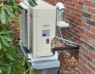 Outdoor air conditioning unit installed on a concrete slab beside a brick wall with connected pipes and nearby green foliage.
