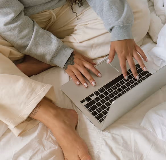 Person sitting on a bed with bare feet, typing on a laptop with both hands visible.