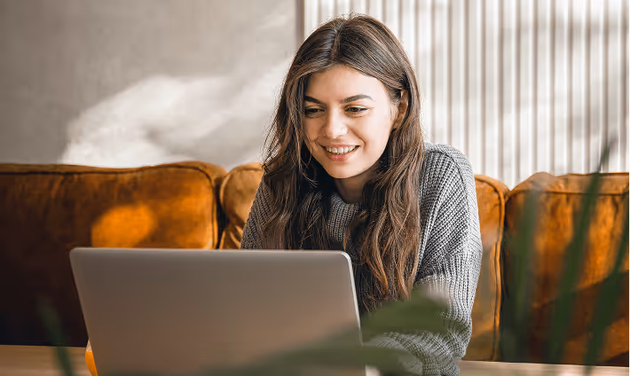 Smiling woman with long brown hair sitting on a brown couch using a laptop.